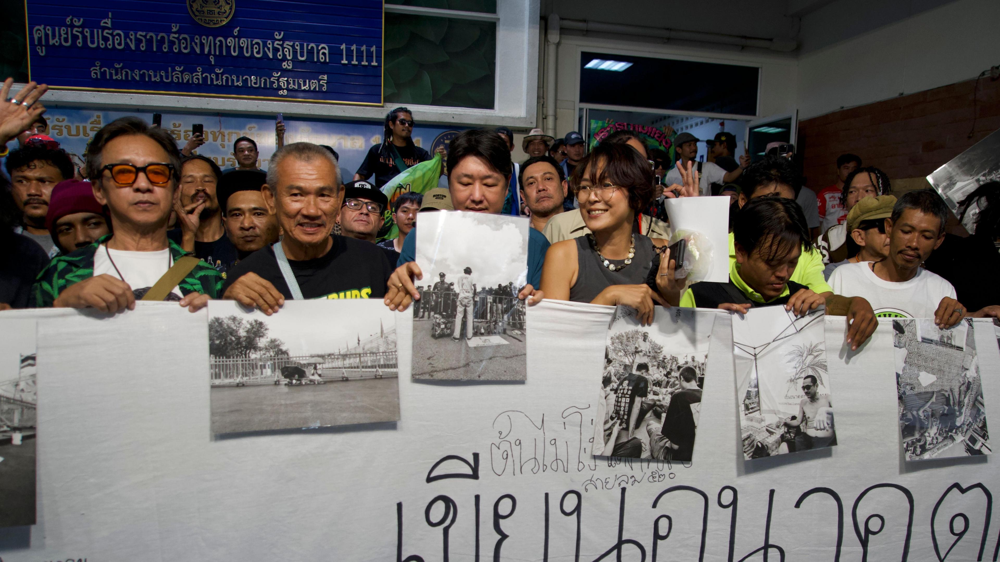 Dozens of people stand holding up a big white banner with black and white pictures attached to it