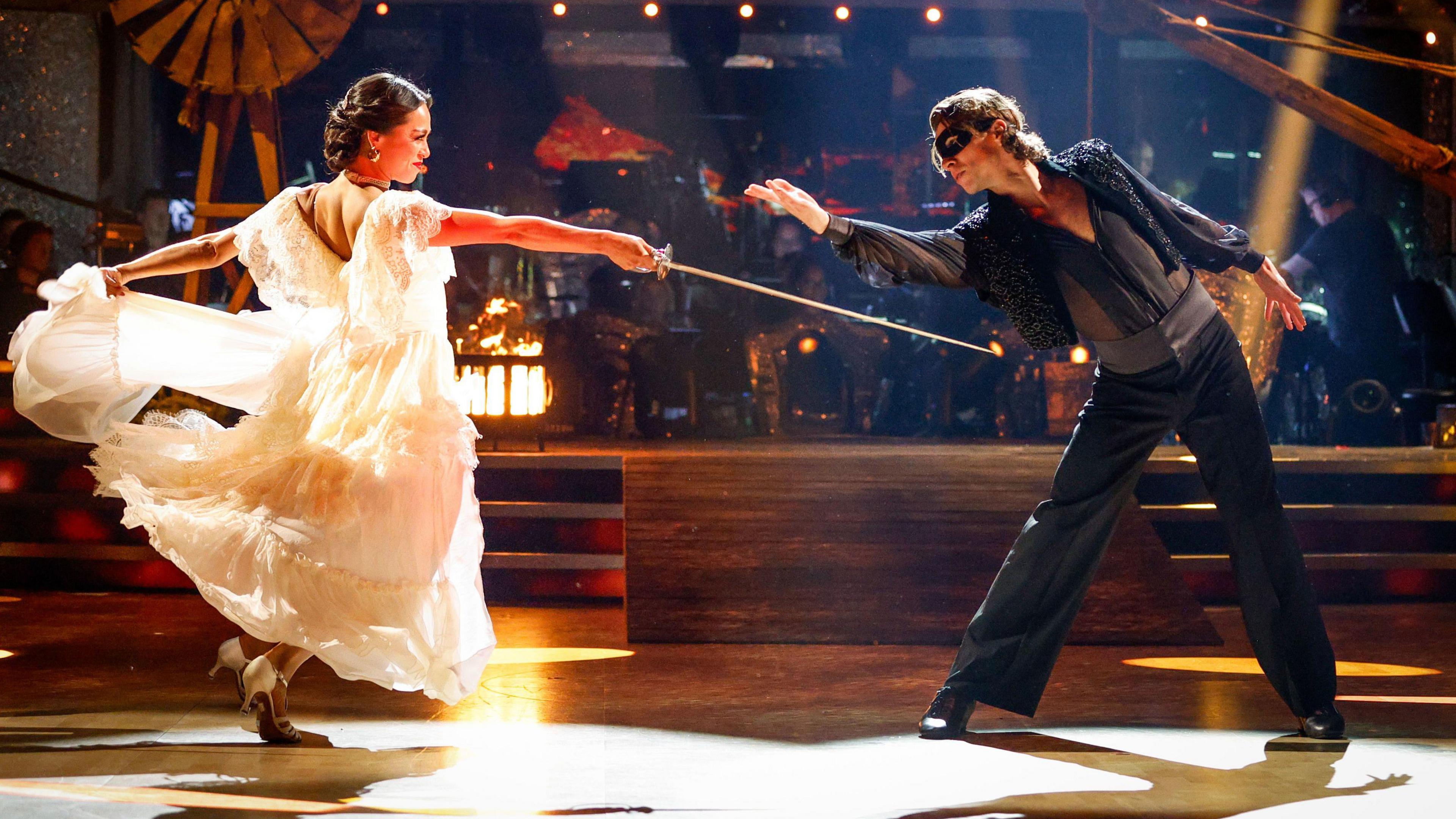 Lewis Cope is wearing black clothes and reaching his arm out whilst wearing a black eye mask. Partner Katya is wearing a flowing white dress and is dancing towards him.
