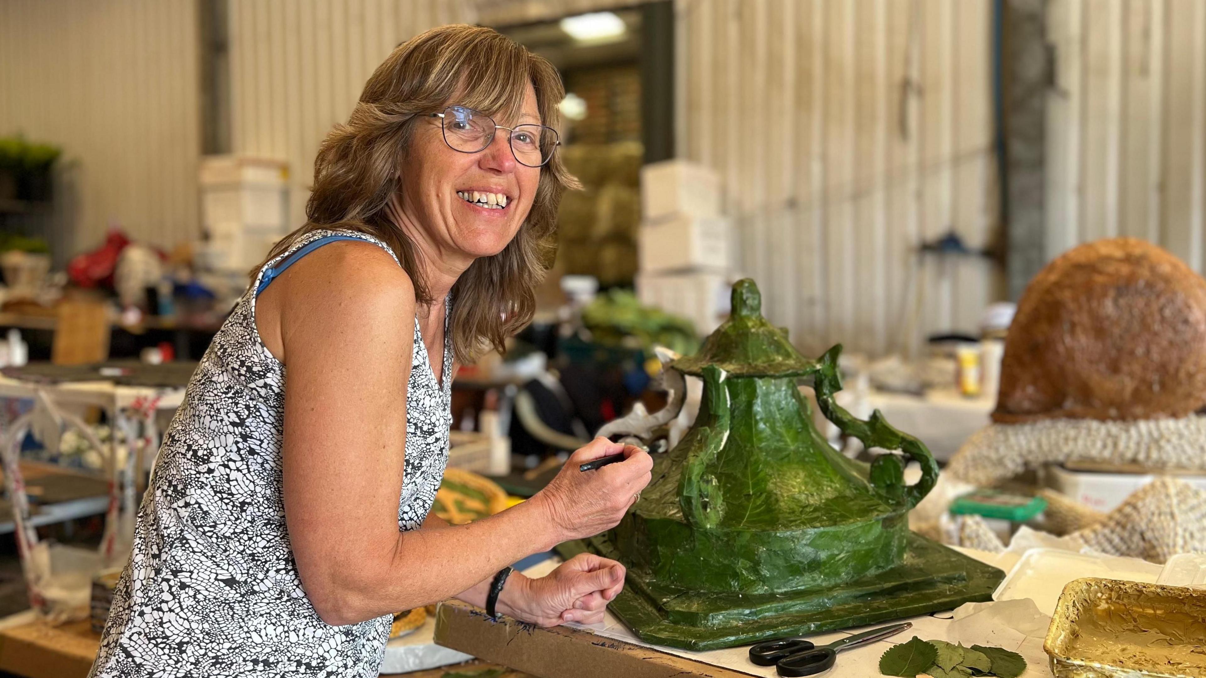 Julie smiling at the camera as she glues leaves on a paper mache lantern. She's wearing glasses and a black and white tank top. Out of focus behind her is a white potato shed with boxes, tables of tools and flowers