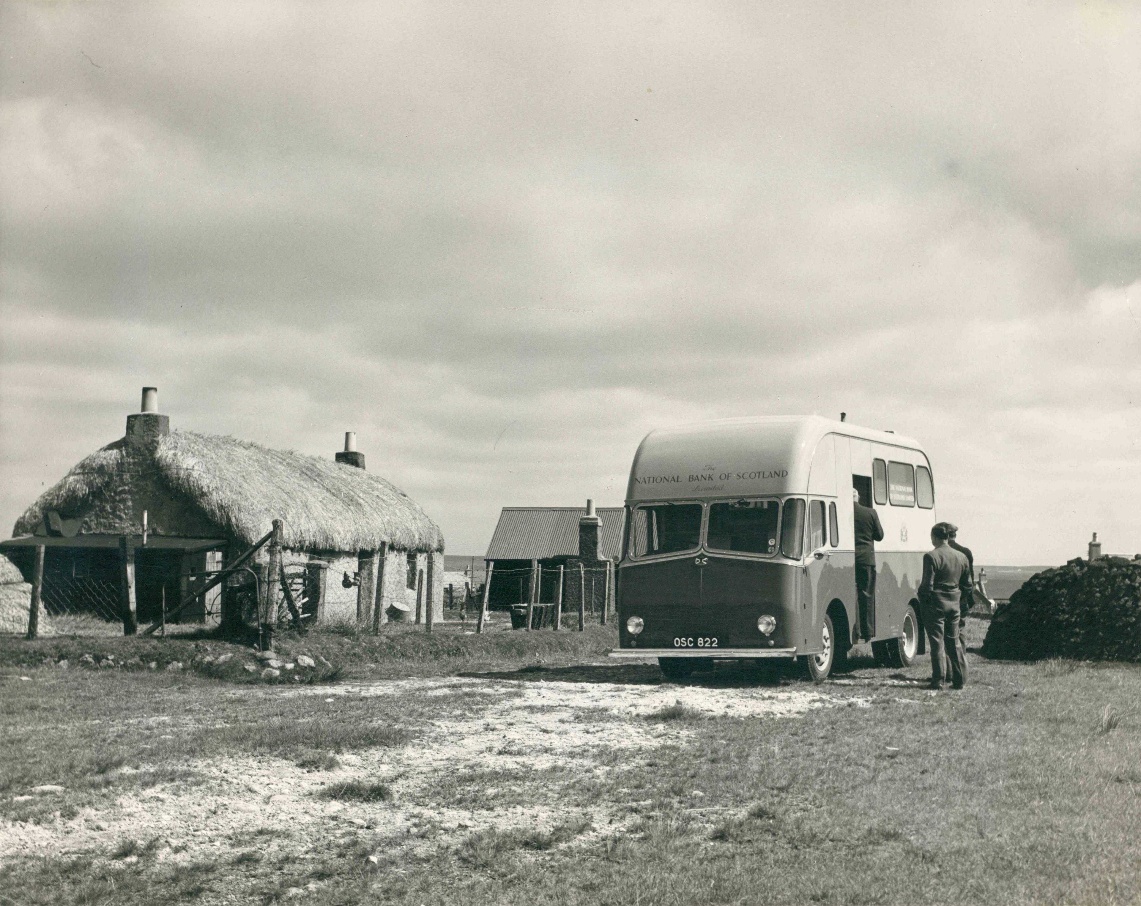 Dealbh dubh is geal de bhana Banca Rìoghail na h-Alba air talamh machrach ri taobh taigh-tughaidh. Tha dithis fhear nan seasamh ri thaobh, le cruach mhònach ri an taobhsan, agus tha aon fhear ann an doras a' charbaid. 