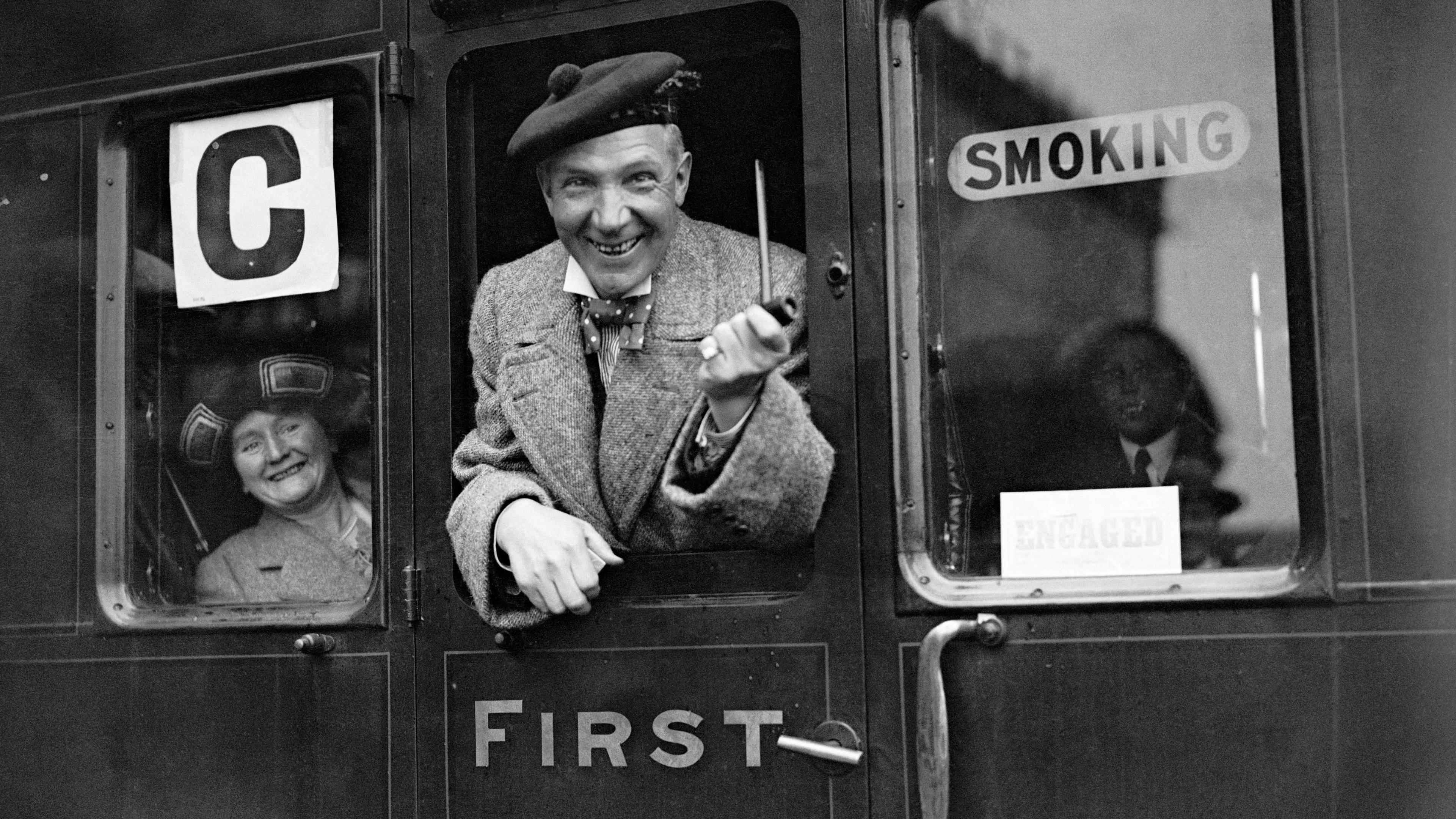 Sir Harry Lauder, a very jolly elderly man with a pipe and lowland bunnet, is hanging out the window of a 1920s railway carriage. A woman in a broad hat and tweed coat is smiling at his antics from inside the train carriage.