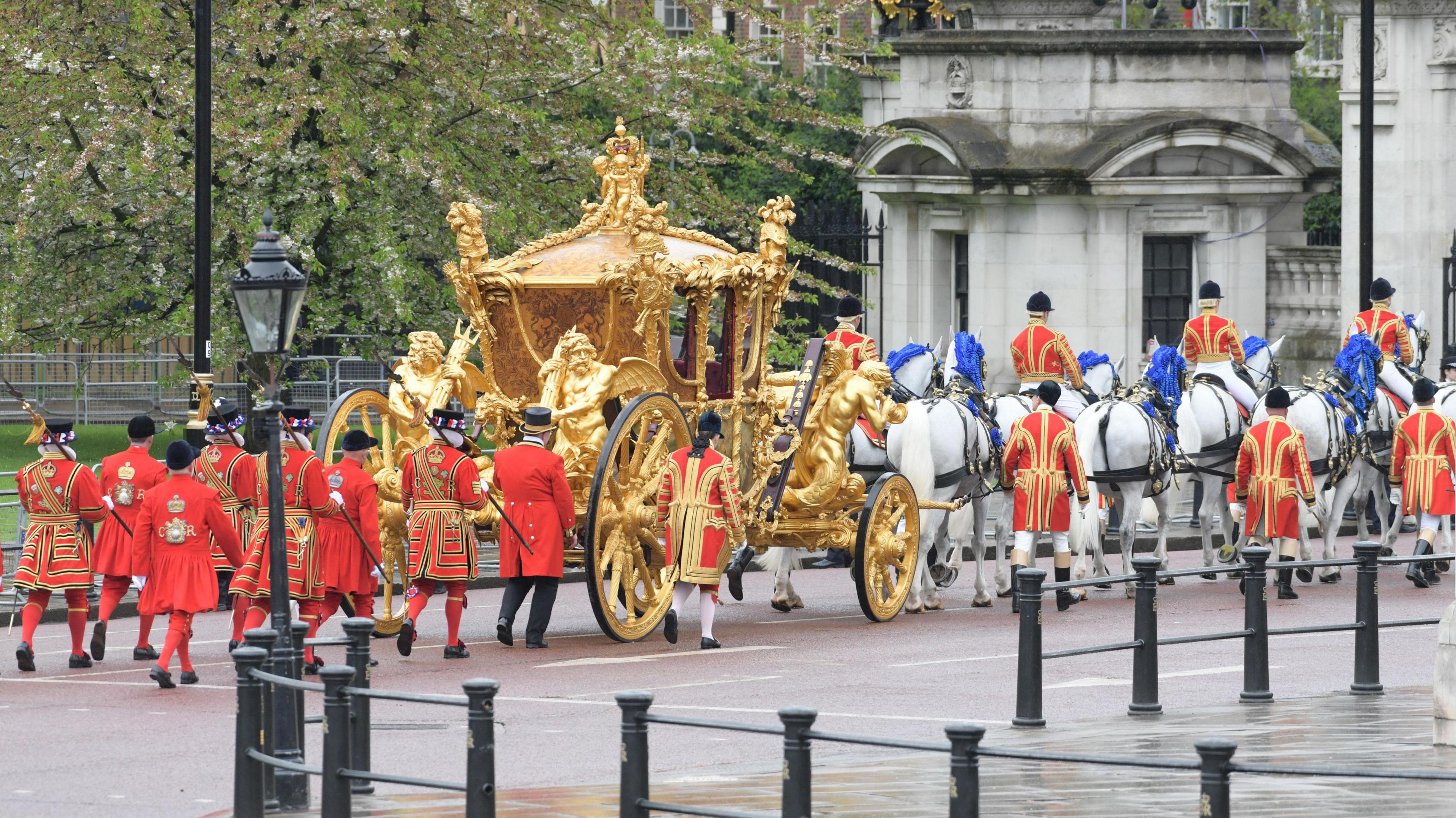 A ornate gold coach pulled by horses on a London street. There are royal attendants in red uniforms following the coach. 