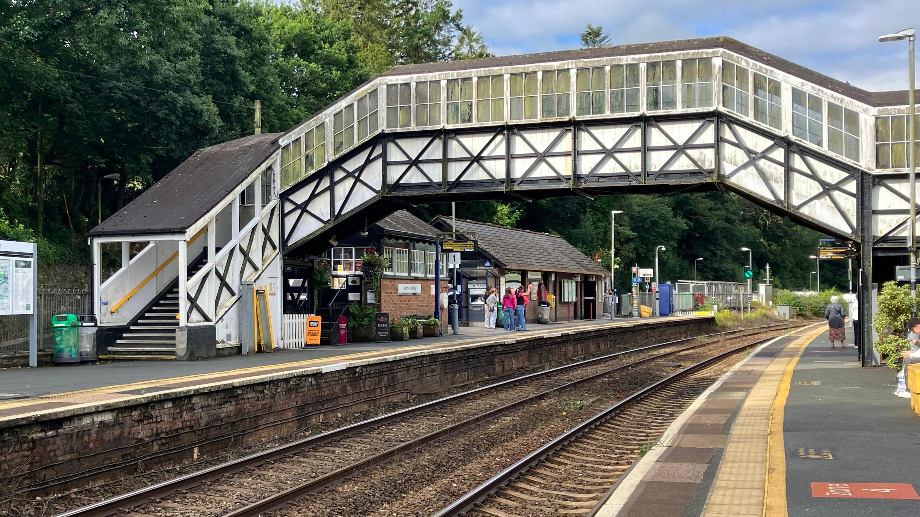 Looking westbound down the platform at Bodmin Parkway at the black and white timber footbridge straddling the tracks underneath.