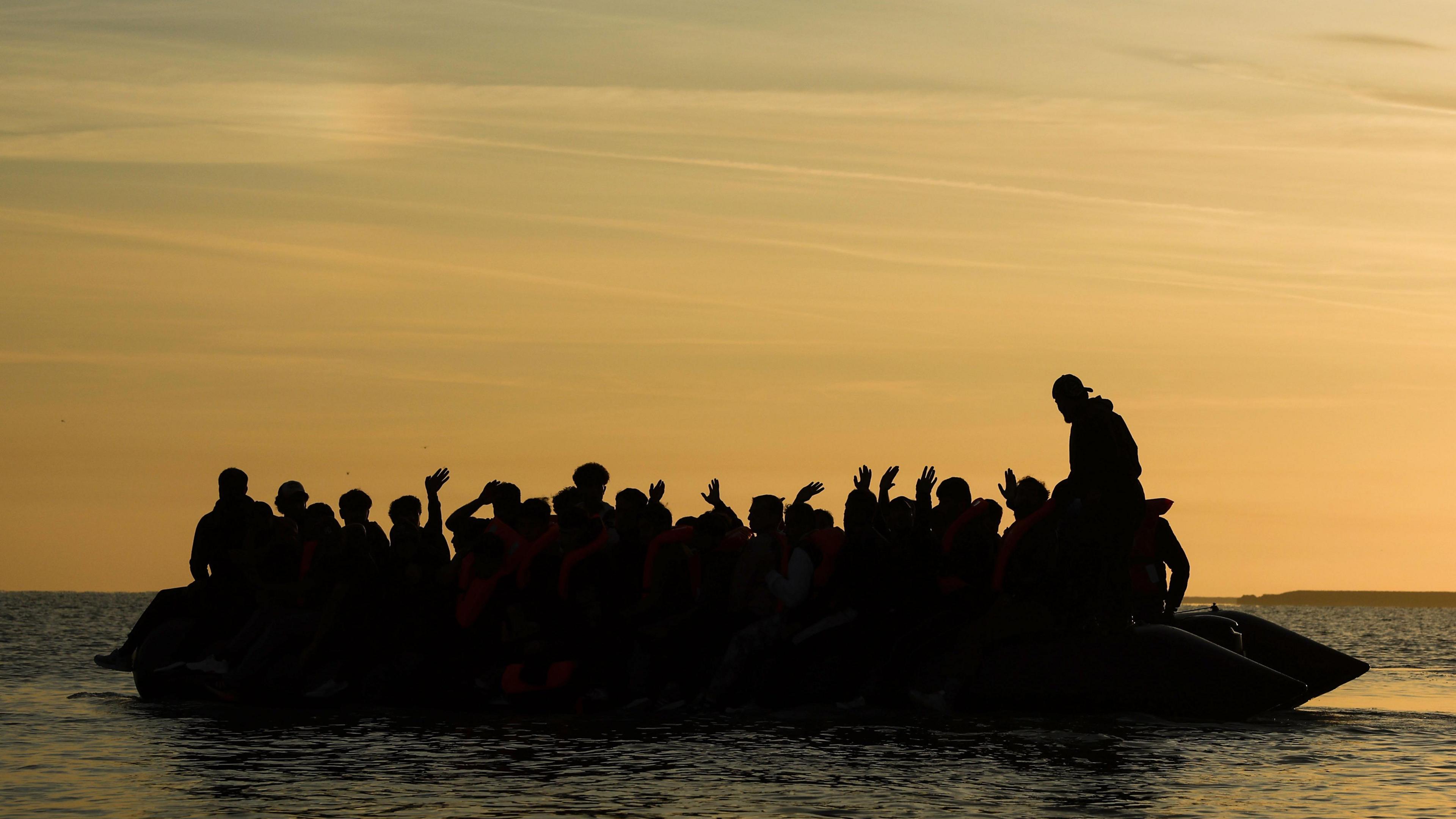 A silhouette image of at least 20 people sitting on an inflatable dinghy at sea. 