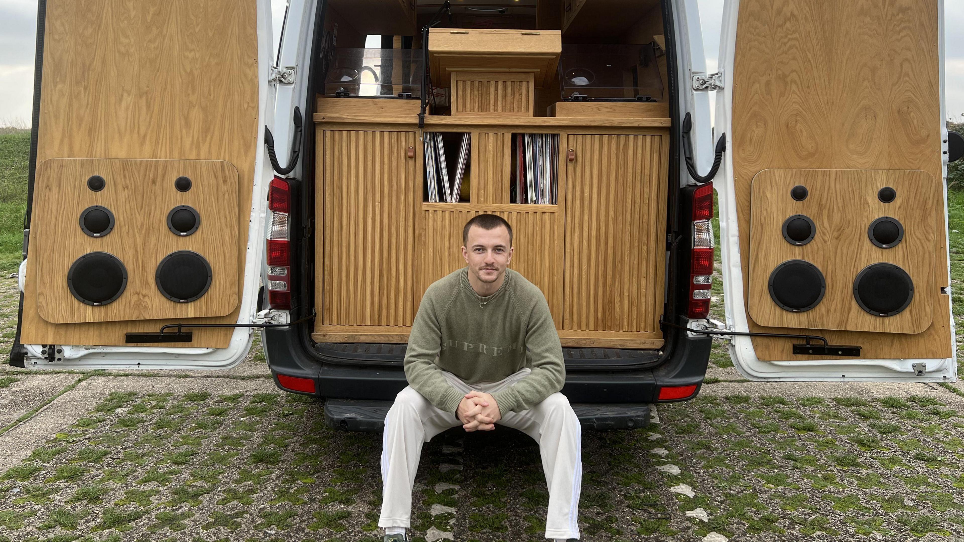 A smiling Jack Lydon sits at the edge of his van, with the doors open and showing his DJ both. My Lydon is wearing white trousers and a green jumper, with his hands clasped together. Behind him you can see speakers attached to the inside of the van's doors and Japanese-inspired slatted cabinetry. 