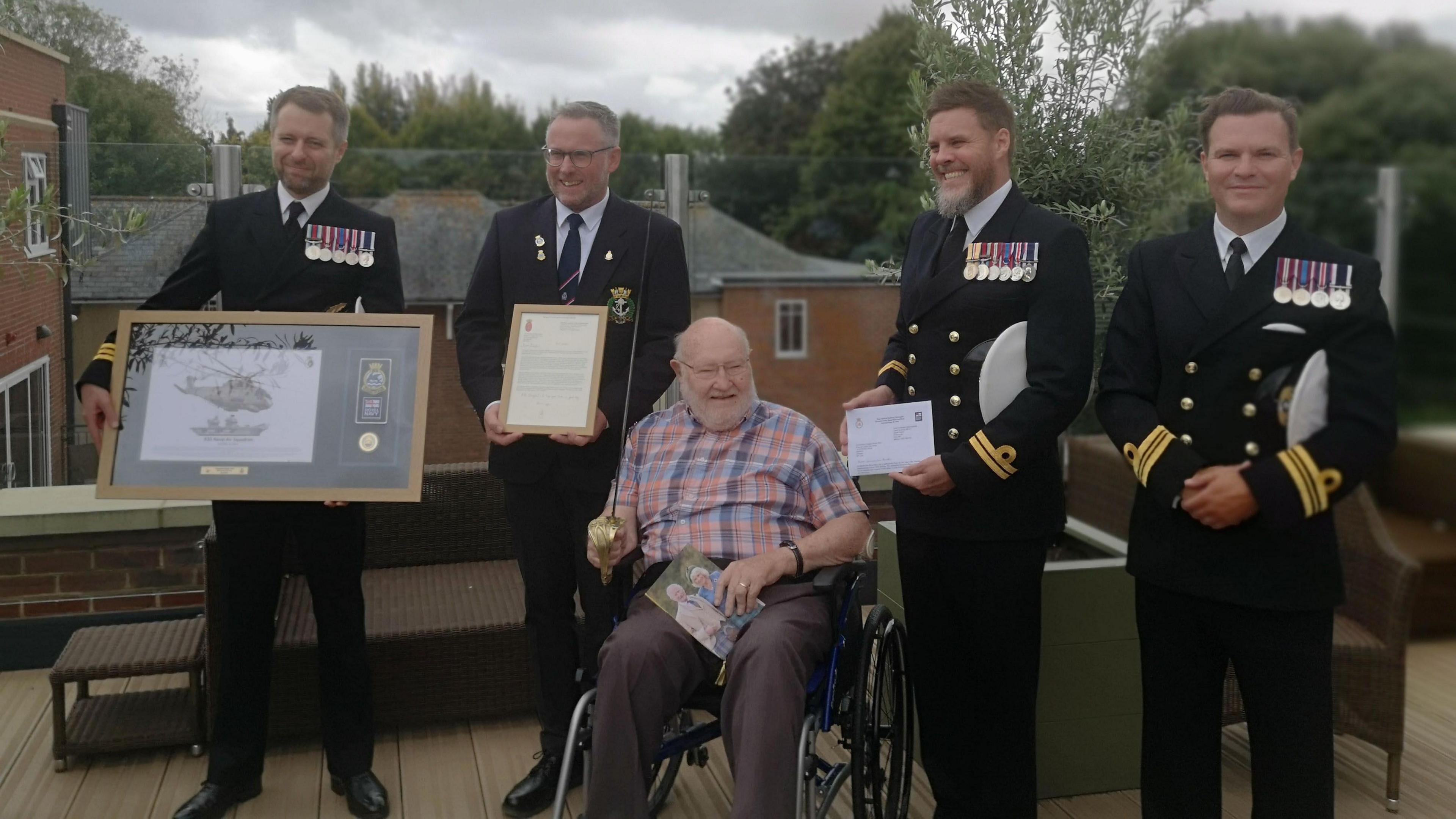 A group of men in naval dress uniform with medals, three holding certificates, one with a pictures. They stand around a man in a wheelchair who is holding a ceremonial sword