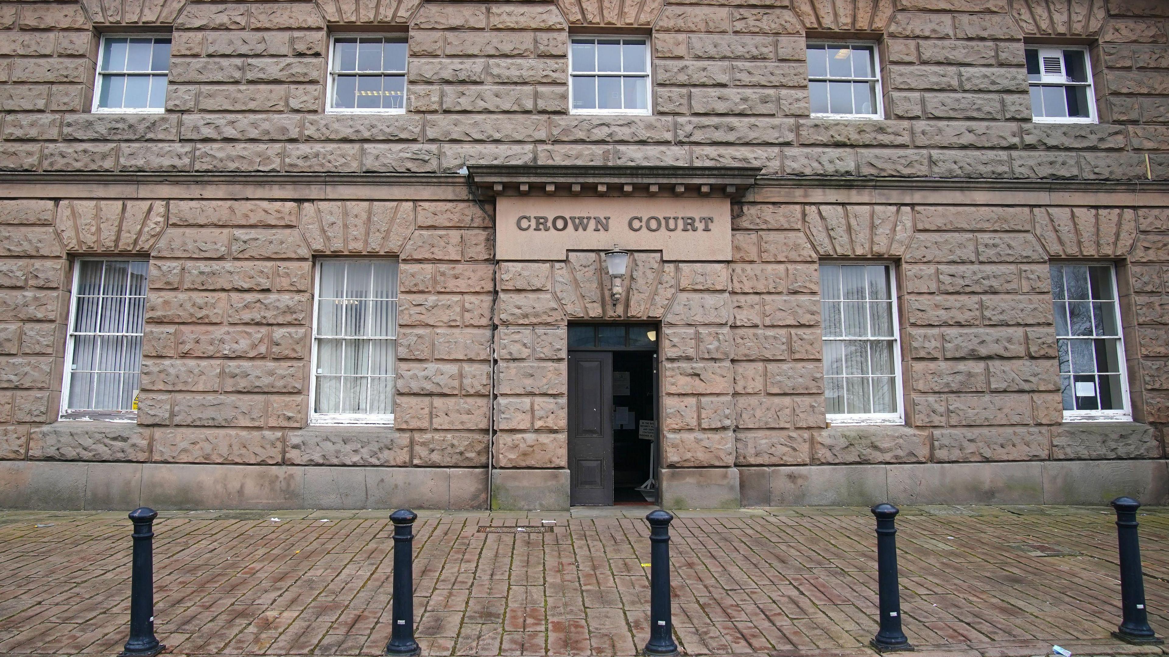 A street view of Chester Crown Court from the roadway. The court is a brown brick building with a black entrance door seen ajar in the shot. Black bollards can also be on the street in front. 