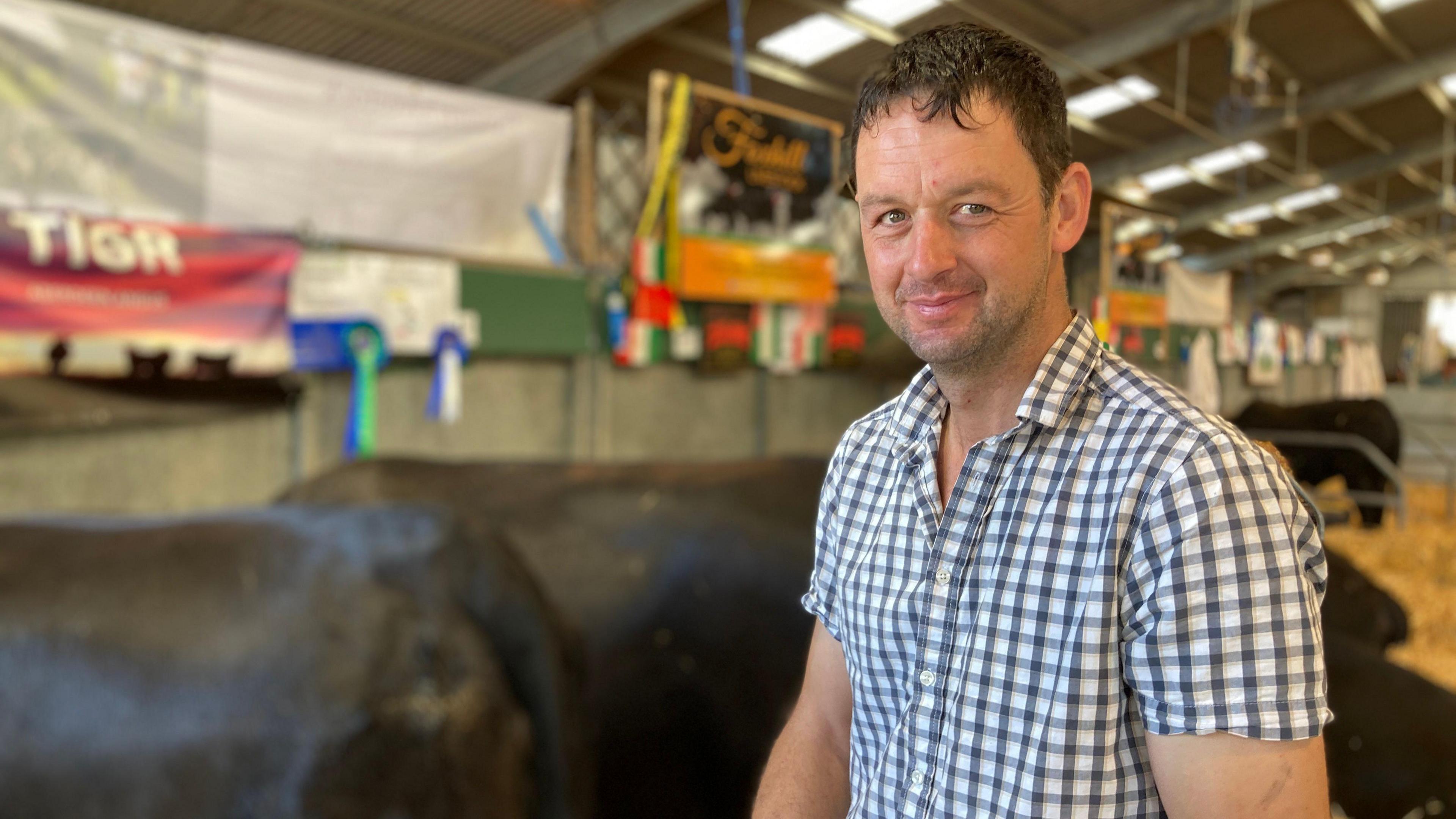 A man with a white and black checked shirt looking at the camera. He has short, dark hair and their are cows behind him.