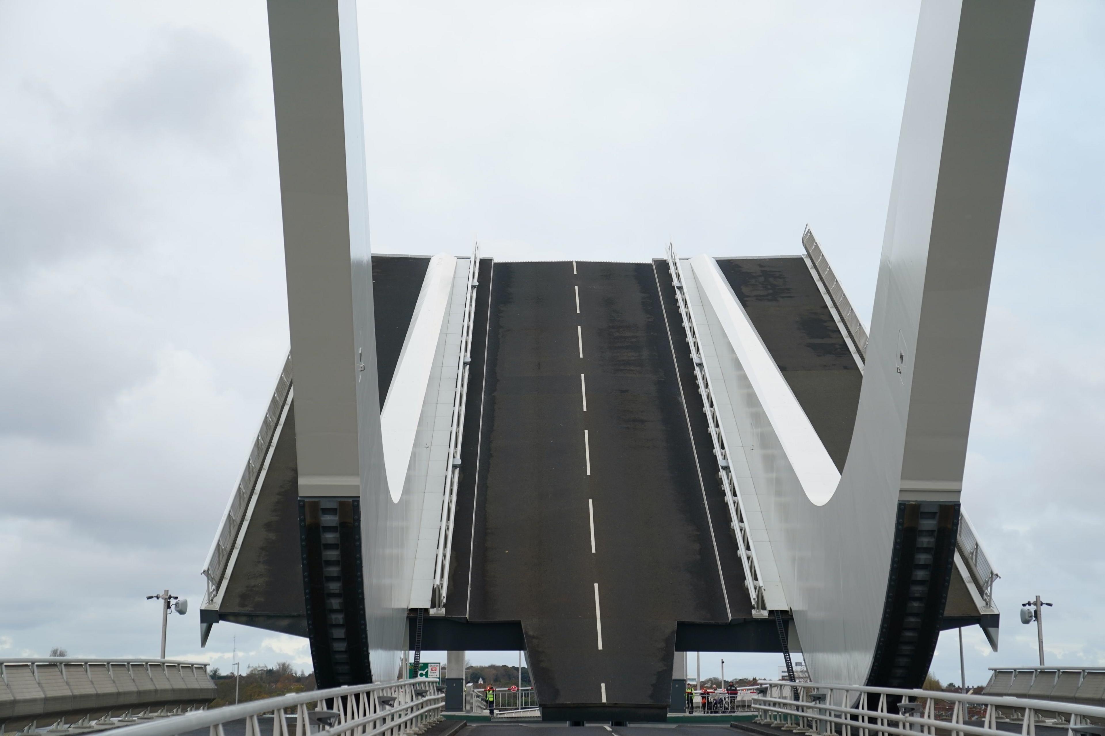 Drone image of Gull Wing Bridge in Lowestoft. It stretches over green river water and has pedestrian walking at either side of the structure. There are two gery curved accents sticking up into the air from the sides of the bridge, almost in the shape of a boomerang. The river can be seen meeting the sea in the background of the image.