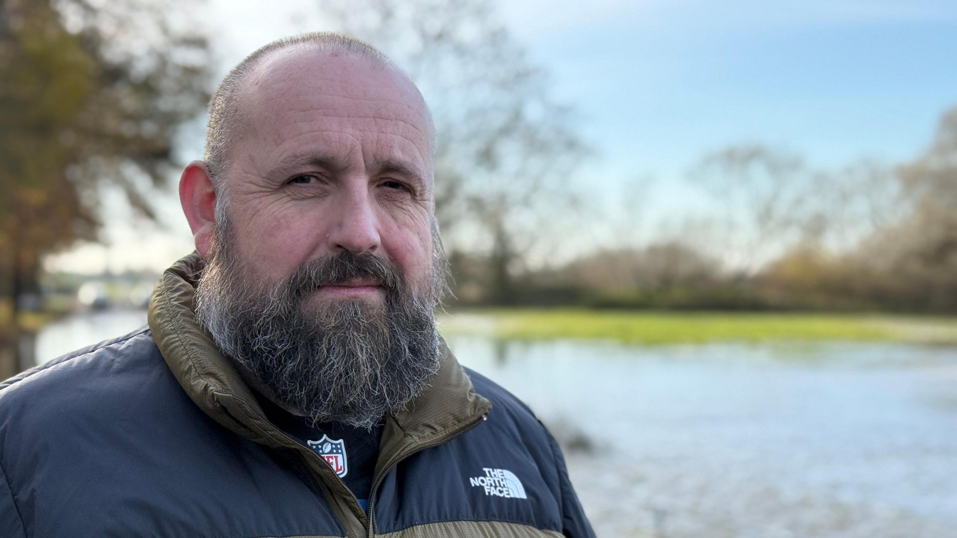 Councillor Stephen Ferguson is looking at the camera with a concerned expression. He has a beard and wears a black and khaki North Face puffer jacket. In the background the flooded road and waterlogged fields can be seen.