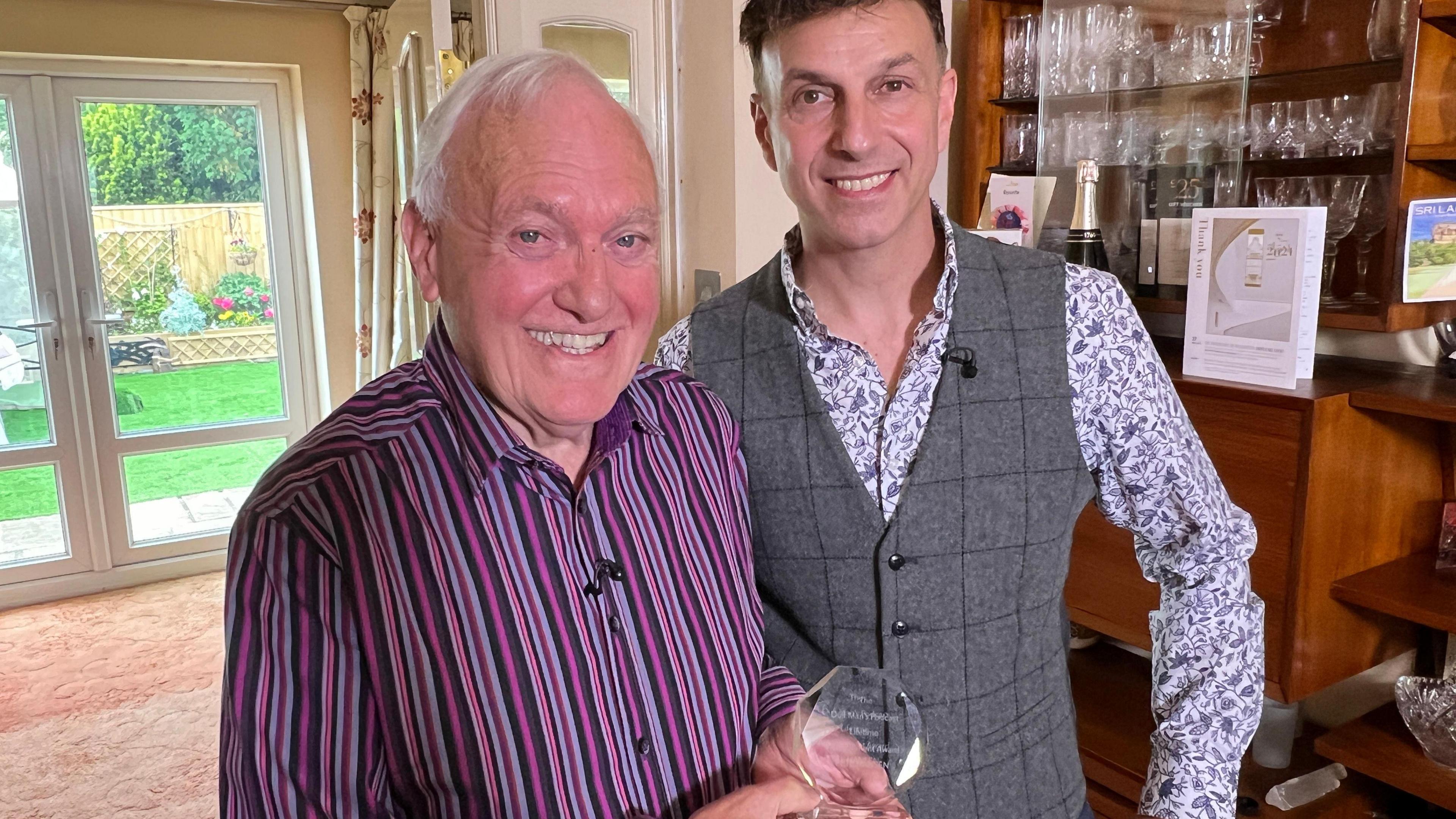 A man is holding a cut glass award standing beside a younger man indoors. Behind them is a glass and wood display cabinet containing various items, and a door opens out to a garden in the background.