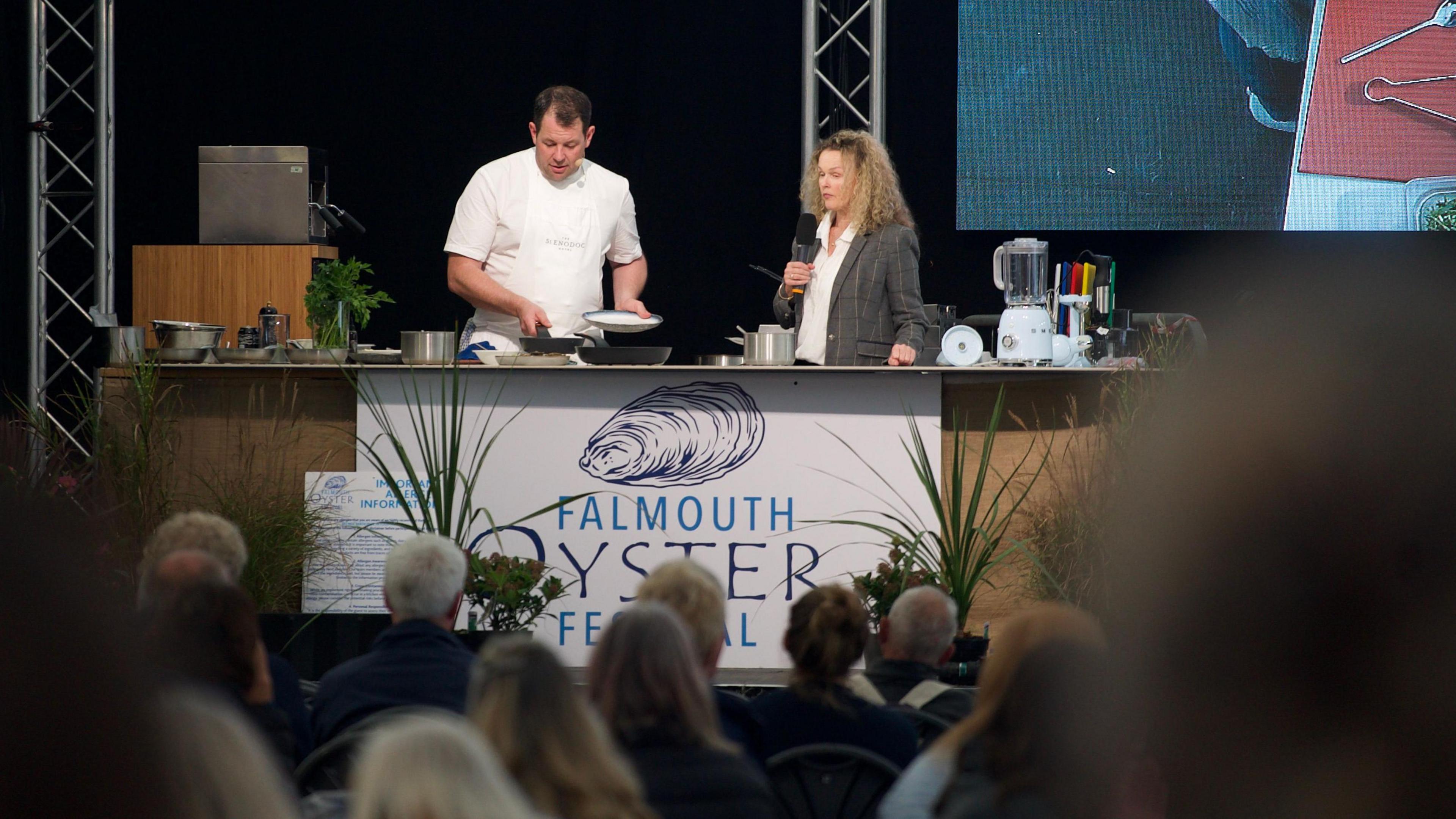 A chef in his whites moves dishes and pans around on a stage while doing a cooking demonstration during the Falmouth Oyster Festival. Dozens of people are watching him. A woman with long curly blonde hair is talking into a microphone on the stage while the chef cooks.