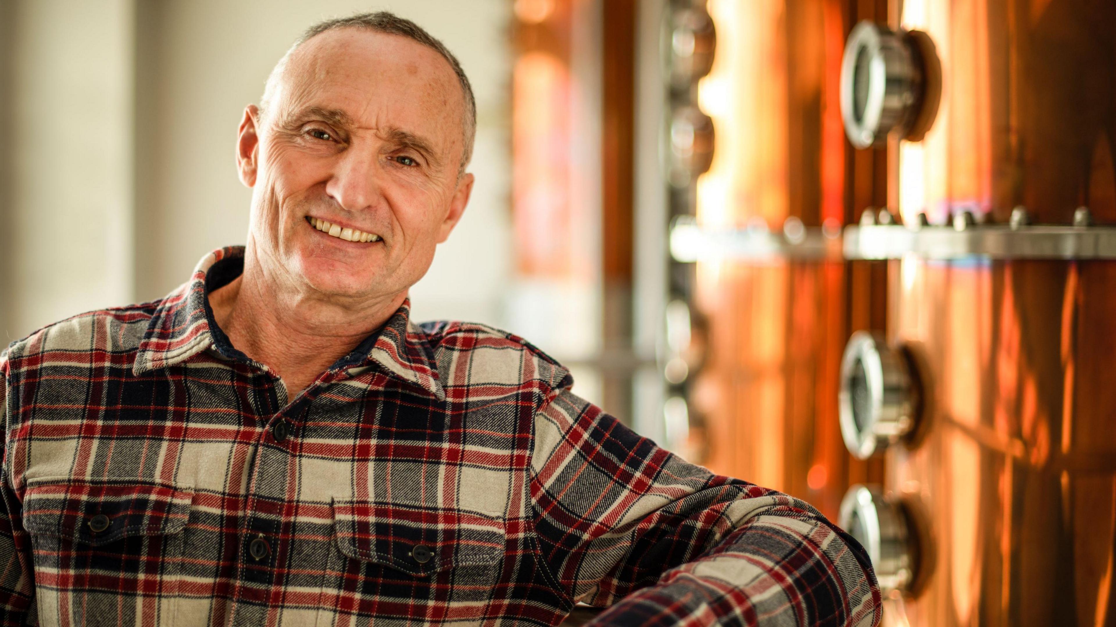 Jonathan Adnams, standing by a beer vat, wearing a checked shirt and smiling at the camera