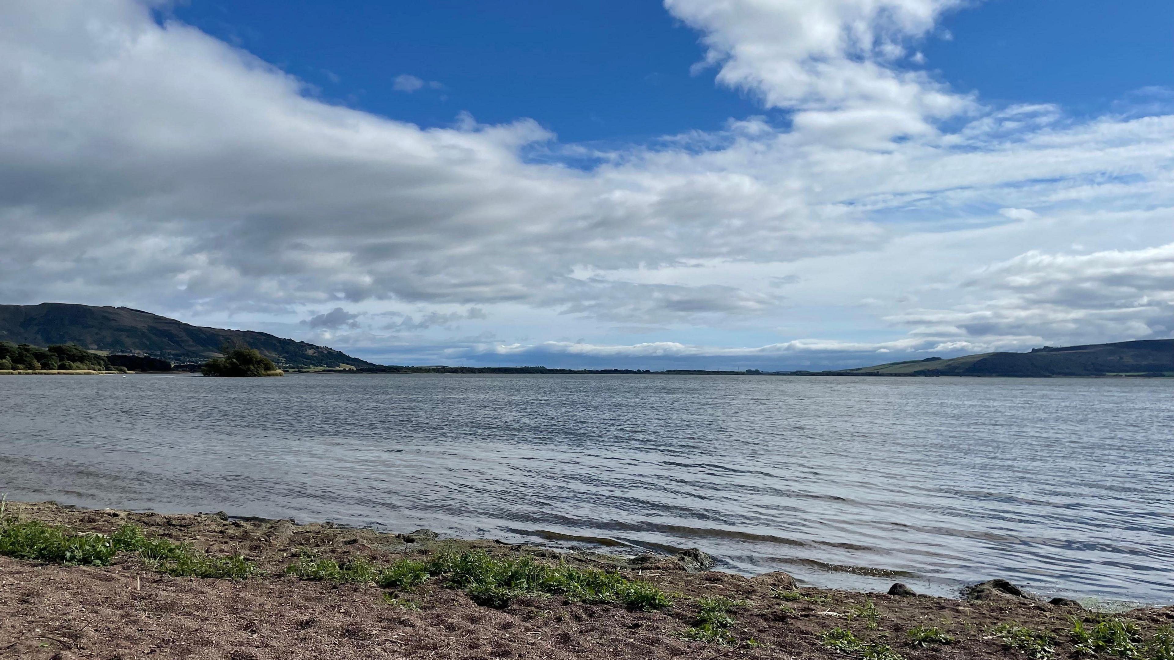 The image show a large body of water surrounded by hills and trees. The photo was taken from the shore on a beach made of brown sand and some greenery. The sky is blue with some clouds.