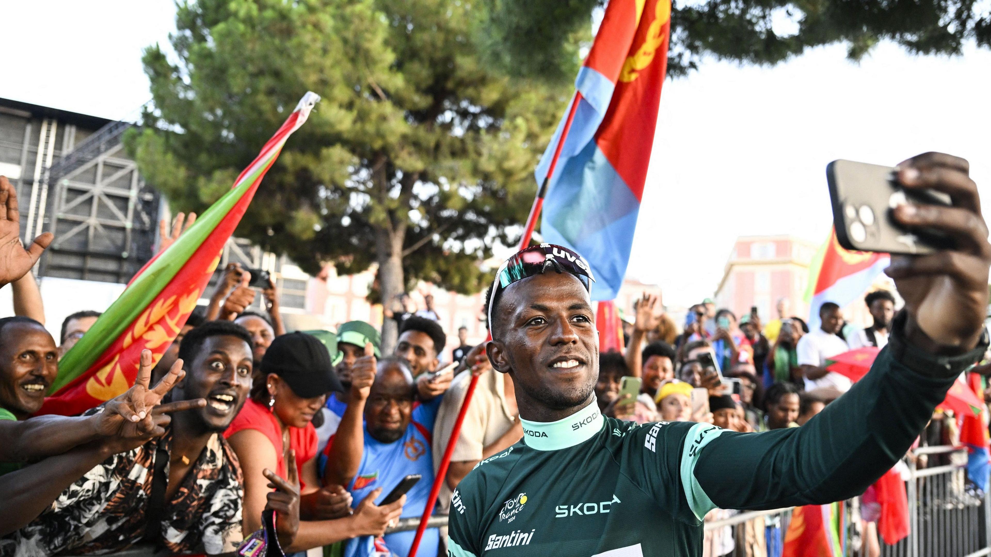 Biniam Girmay seen from chest up, wearing a green cycling jersey, extends his left arm to take a selfie in front of fans behind crash barriers who are waving Eritrean flags. A large tree in leaf can be seen rising in the background 