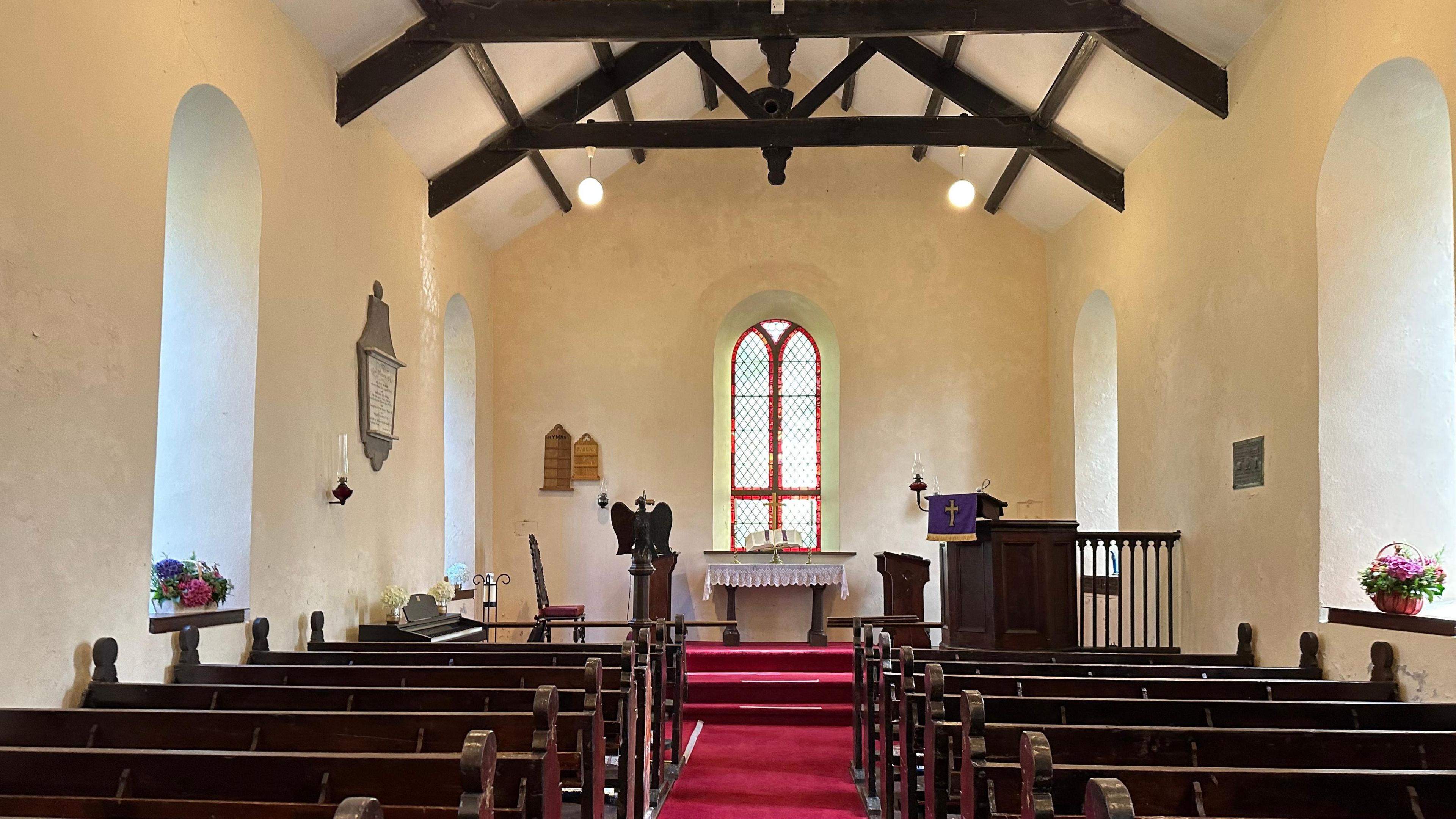 The interior of the small Church, the walls are cream, there are dark wooden pews and the carpet is red, there is a long window at the front and an alter.