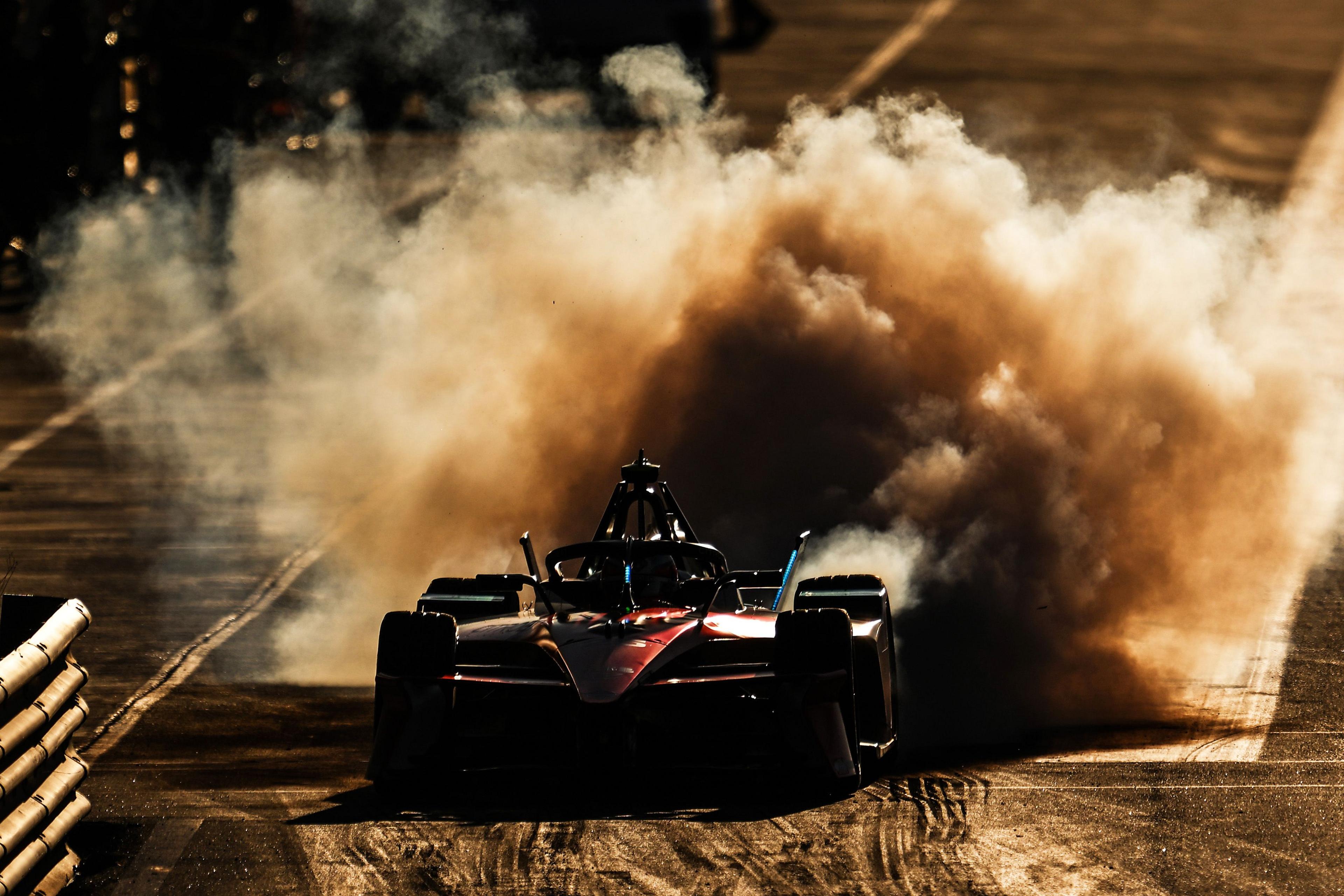 A Formula E race car speeds down a sunlit track, kicking up a massive cloud of dust and smoke behind it. The vehicle’s sleek aerodynamic design contrasts with the gritty surface, creating a dramatic scene of motion and intensity.