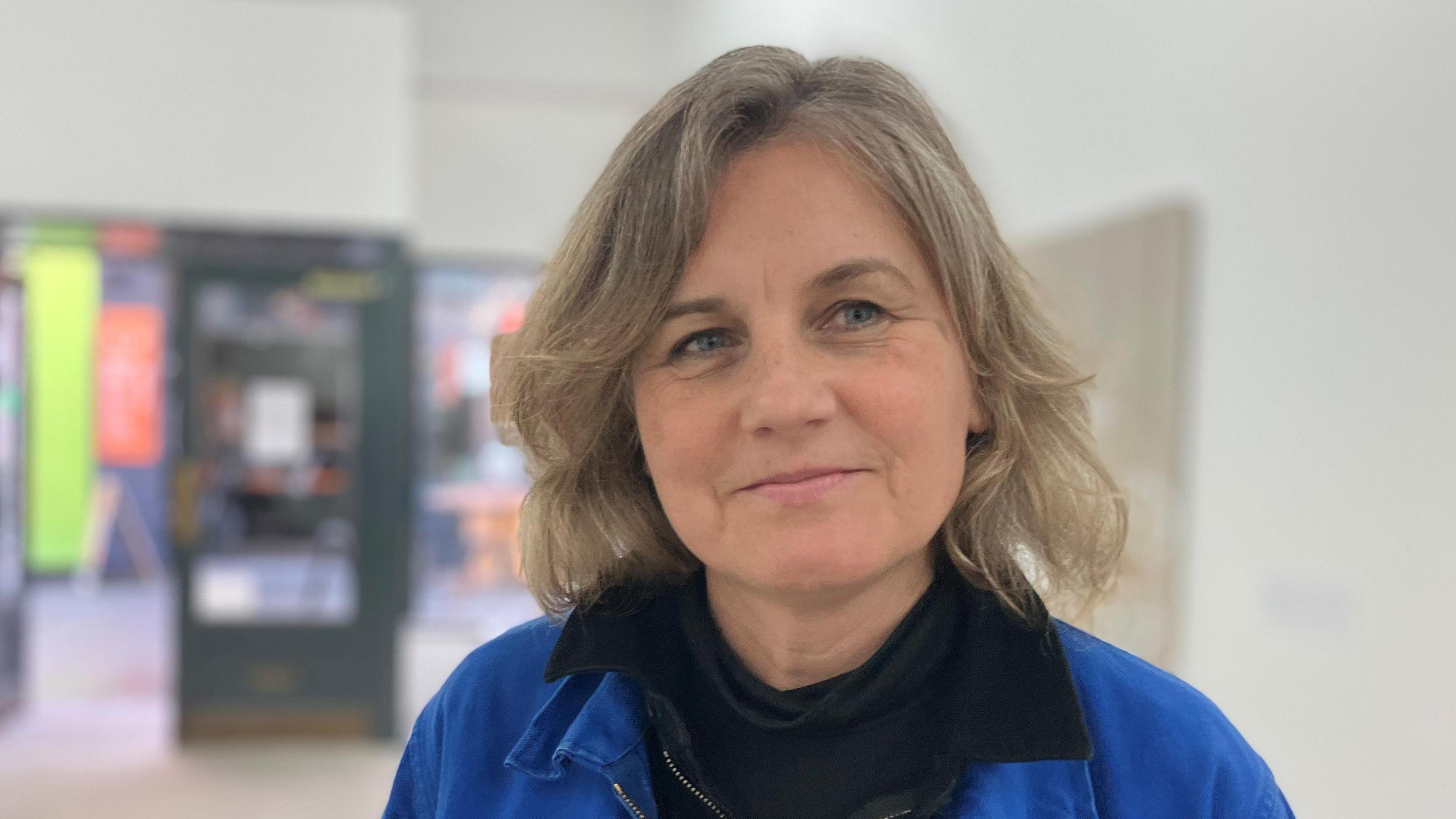 Kate Jones is pictured in a head-and-shoulders image. She has wavy blonde hair in a bob and is wearing a black roll-neck top with a blue sweatshirt over it. She is smiling at the camera and is standing in front of some artwork displayed on a wall.