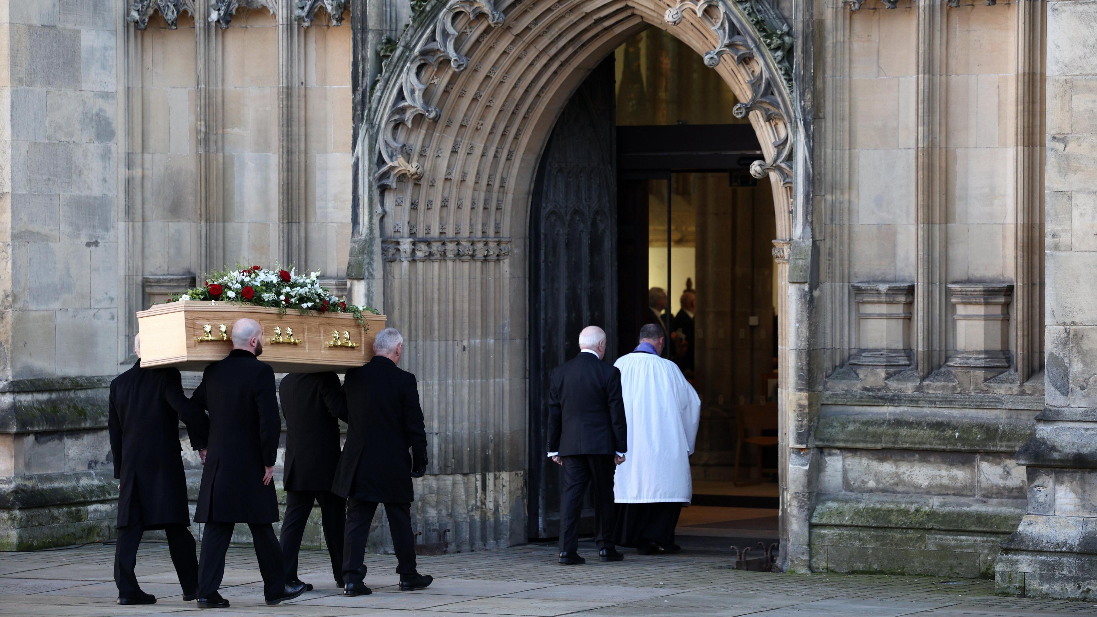 A wooden funeral coffin with flowers on top being carried into Hull Minster by men dressed in black