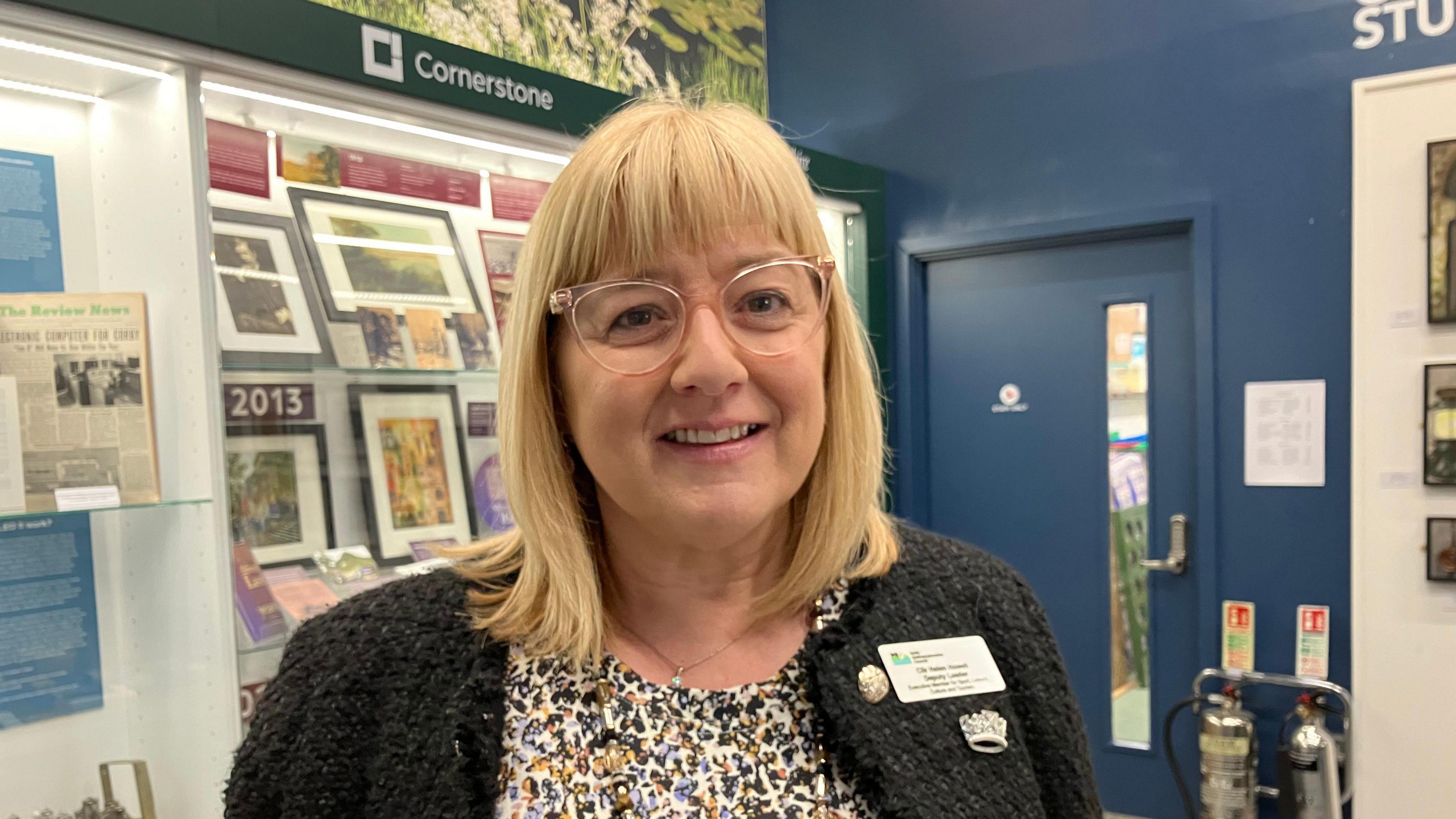 A woman with blonde hair and glasses wearing a charcoal knitted cardigan stands in front of an art display.