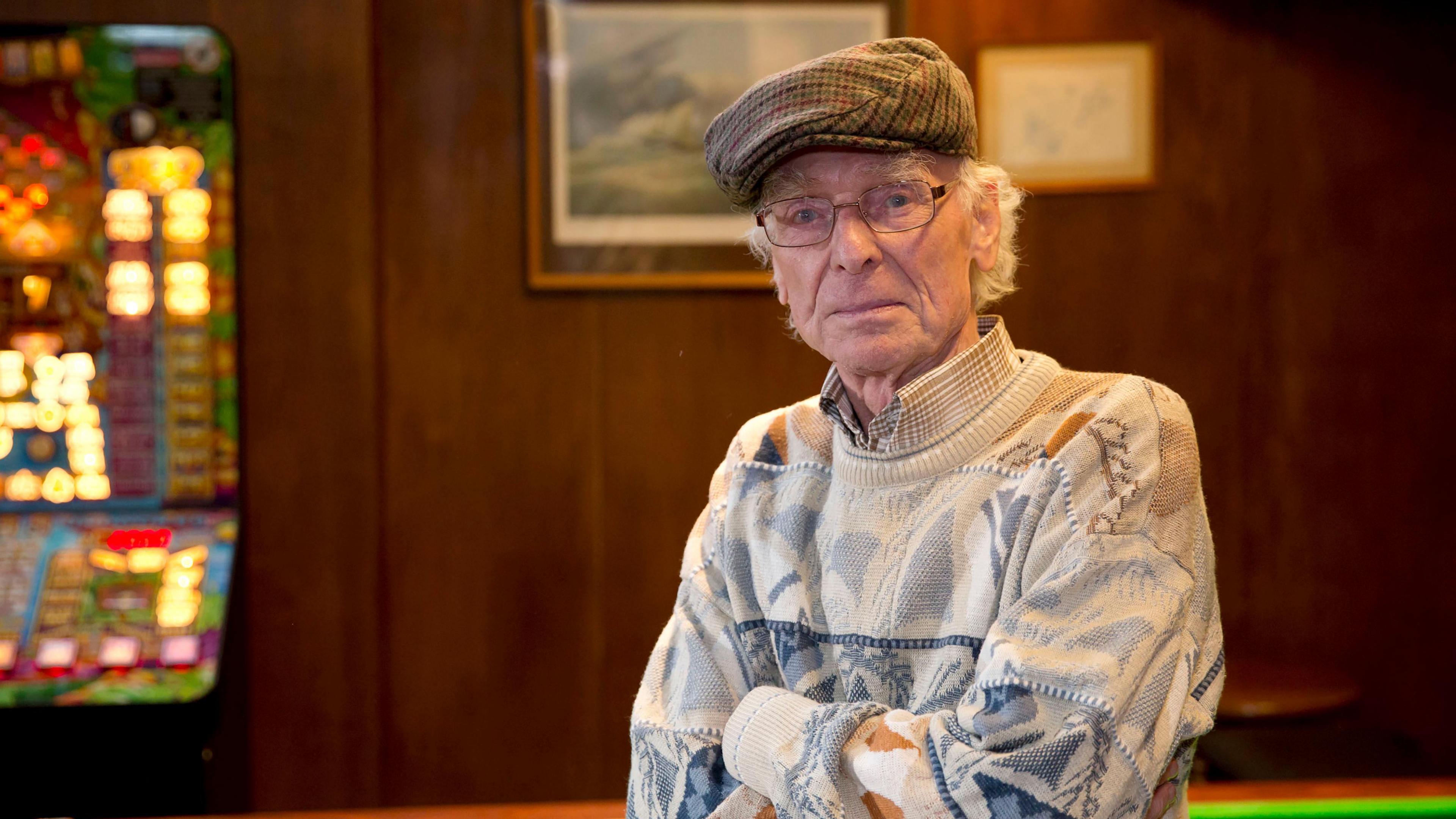 Jimmy Martin looking directly at the camera. He is wearing a multicoloured jumper and has his hands folded across his stomach. He is wearing a multicolured light bunnett and is standing in front of a wood background. A blurred fruit machine is on the right of the picture.