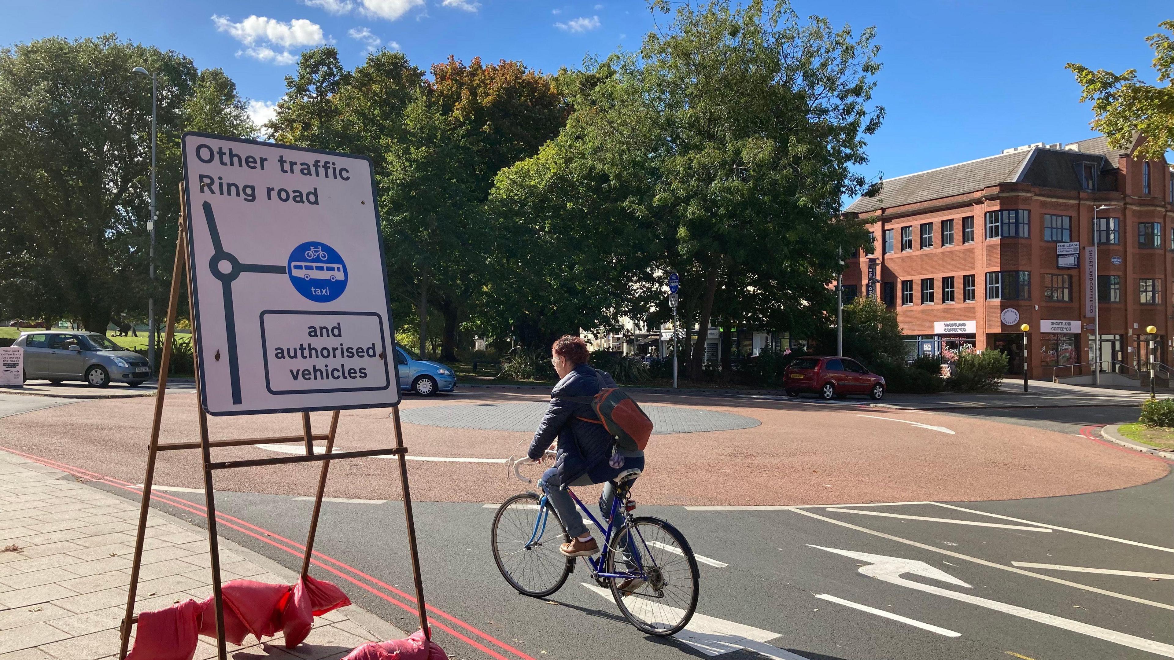 A roadsign with black text and white background with other traffic and ringroad written on it and authorised vehicles is on a stand next to a traffic island on a road. There are cars in the background and a cyclist in the foreground