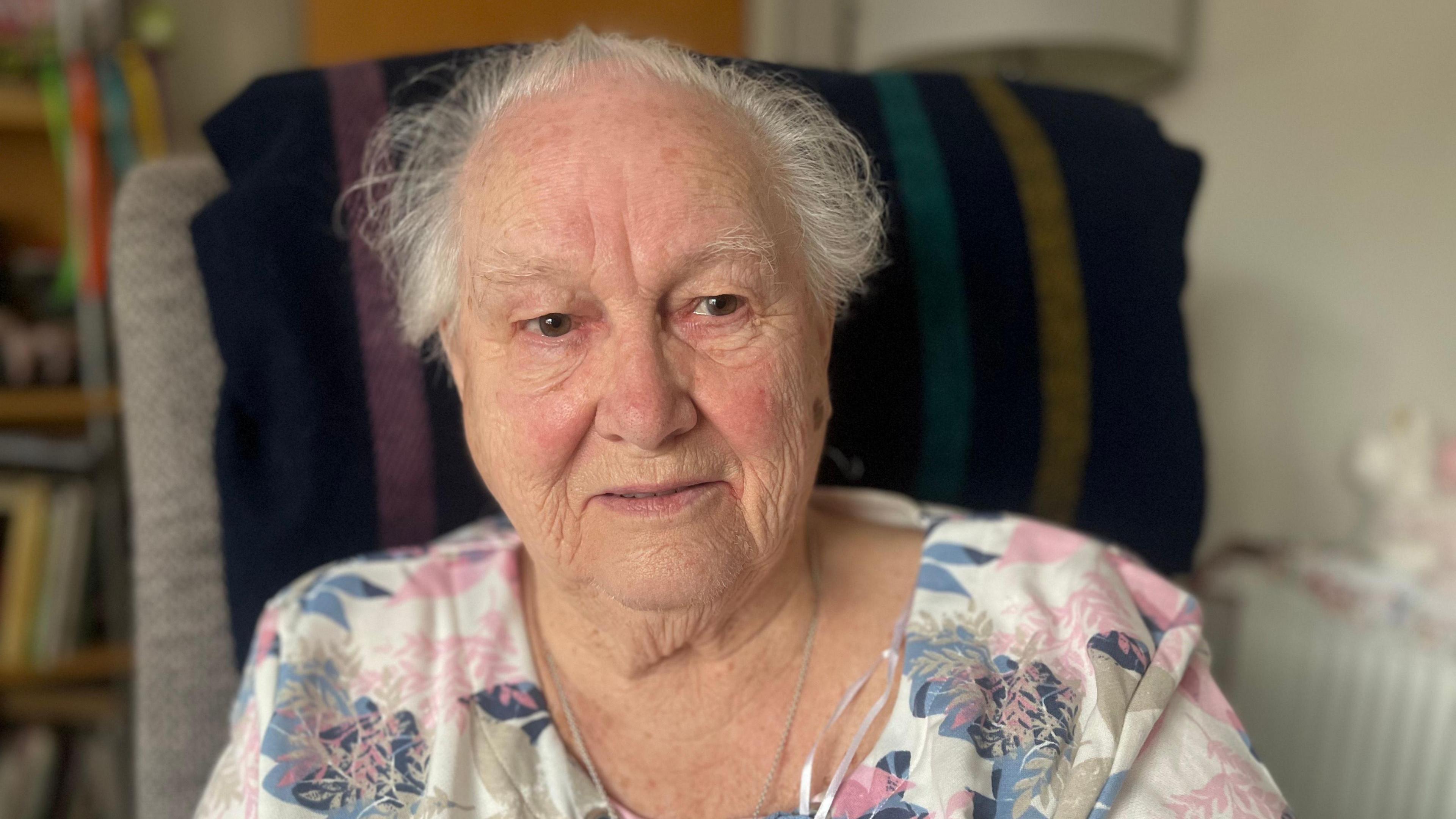 Anne Greenan sitting on a chair, to the right of the frame, with a door and a shelf in the background of the image.