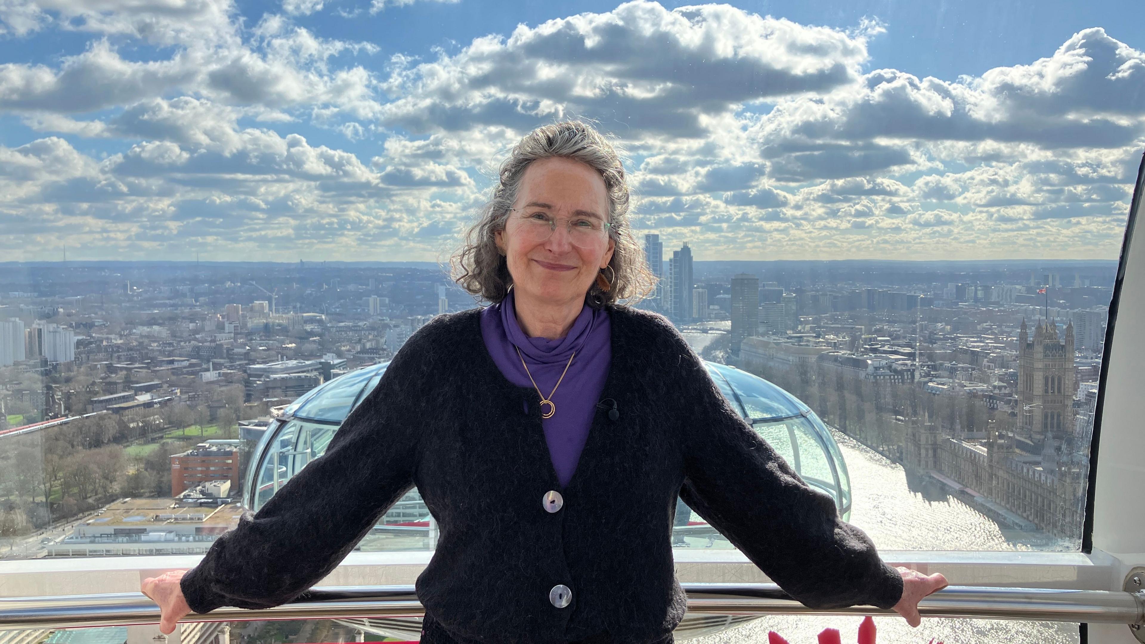 Julia Barfield, a woman with short, wavy grey hair stands inside a capsule on the London Eye; in the background the bright sky is full with clouds and there is a view of the Houses of Parliament