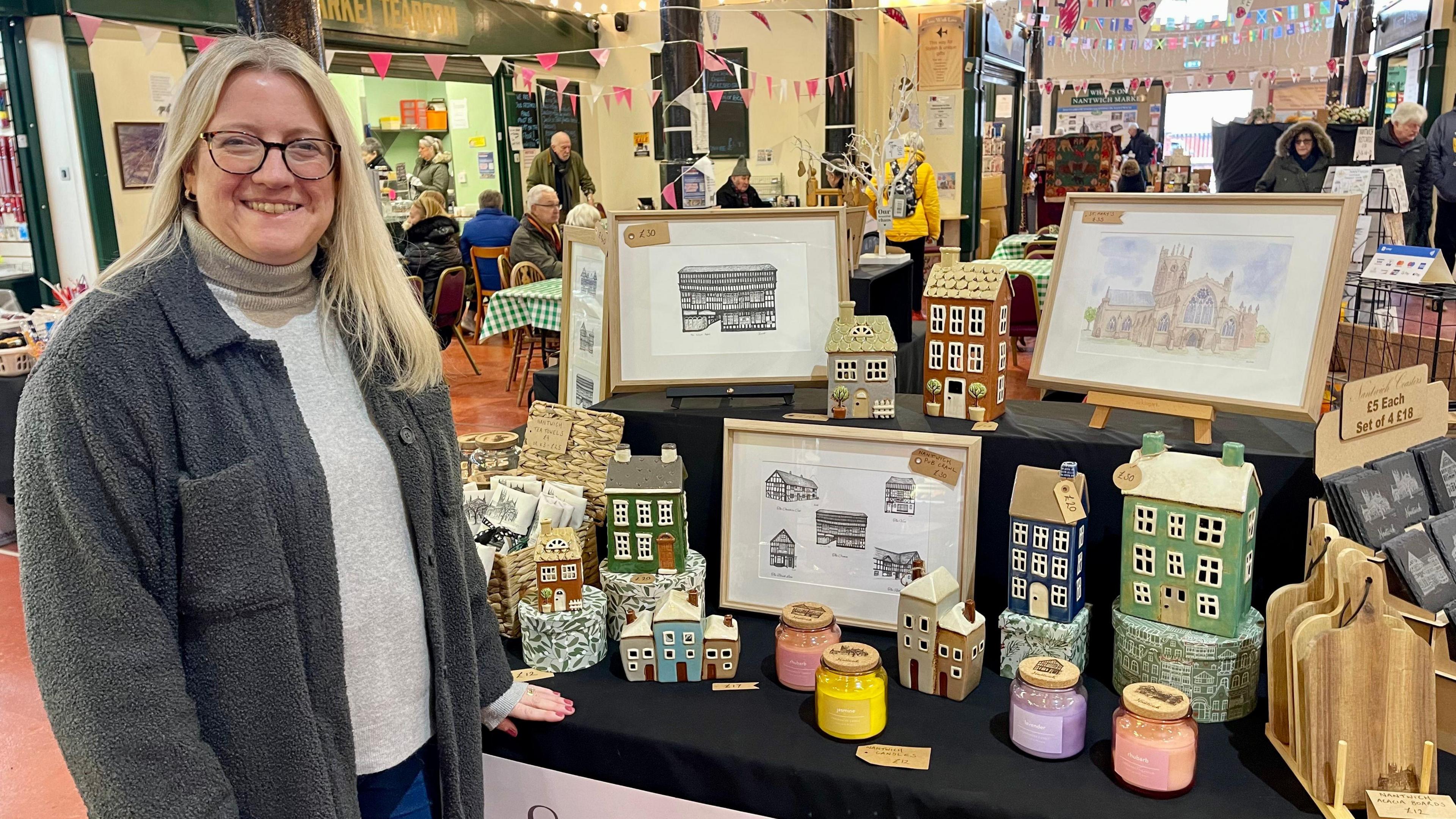 A lady stands by a market stall covered in paintings, drawings and model clay houses. She has long blonde hair, glasses, a grey fleece jacket and a cream jumper. Behind her, people are sat eating and drinking in the market cafe with bunting hanging above their heads.