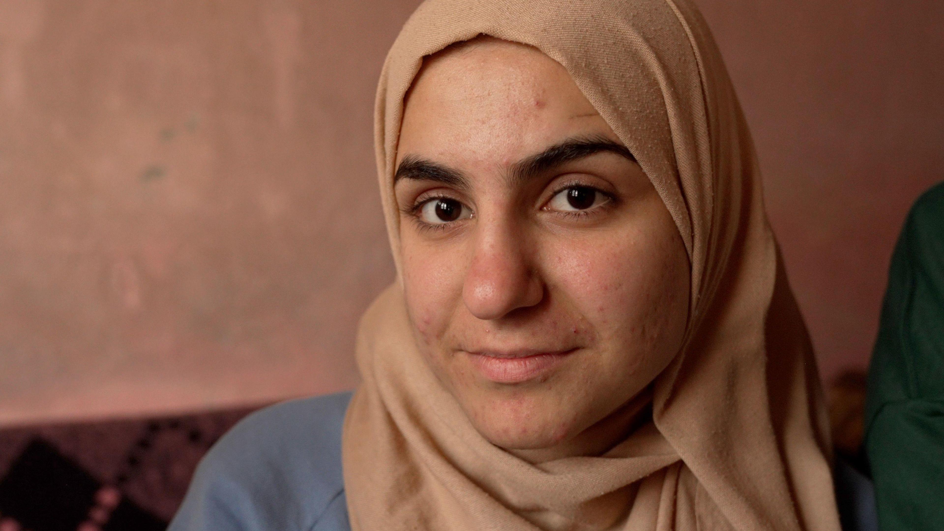 Baraa stares down the barrel of the camera, wearing a beige headscarf, sitting inside a room 