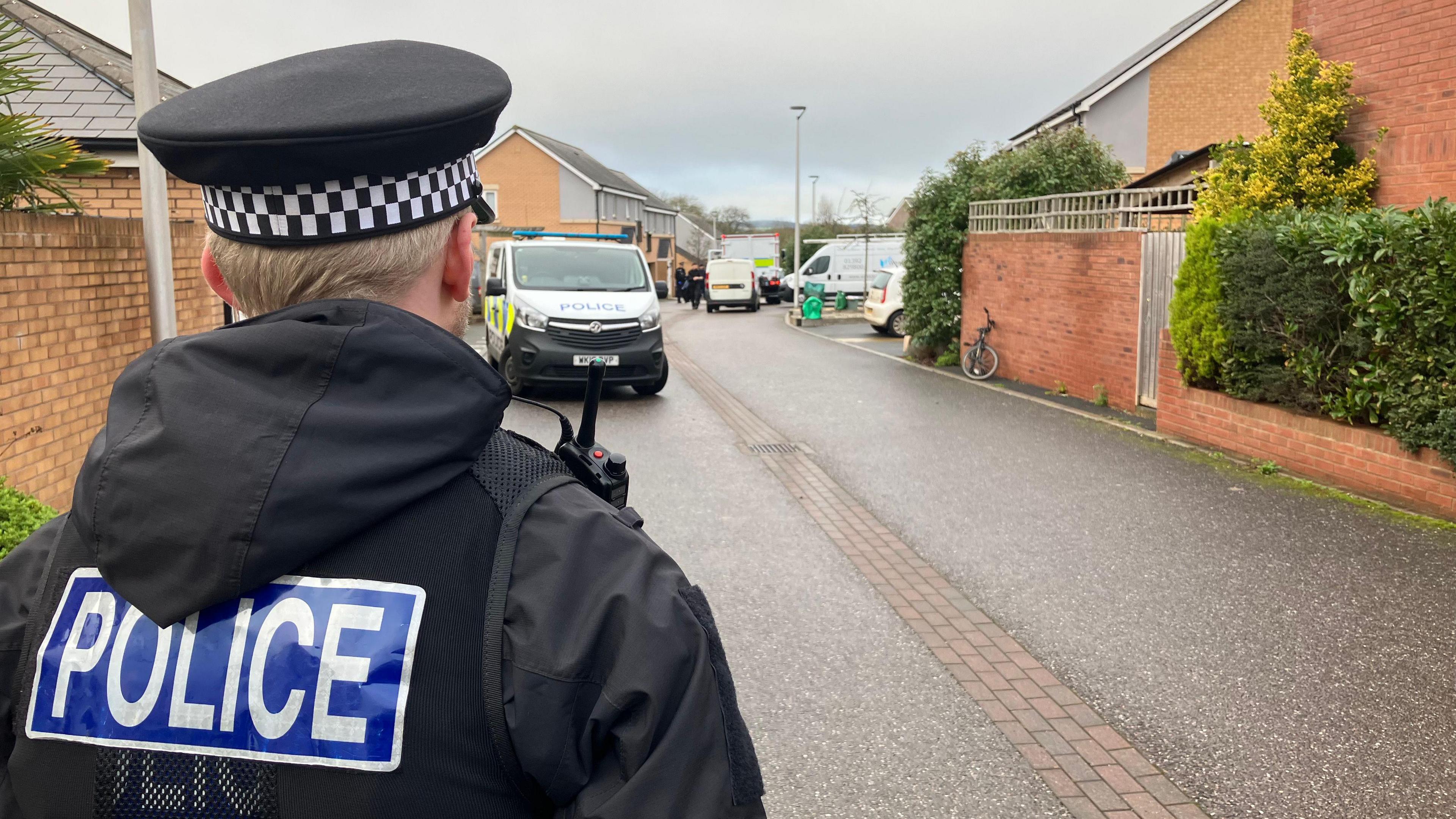 A police officer is photographed from behind as he watches over the scene further down the road where a police van and a large white vehicle can be seen.