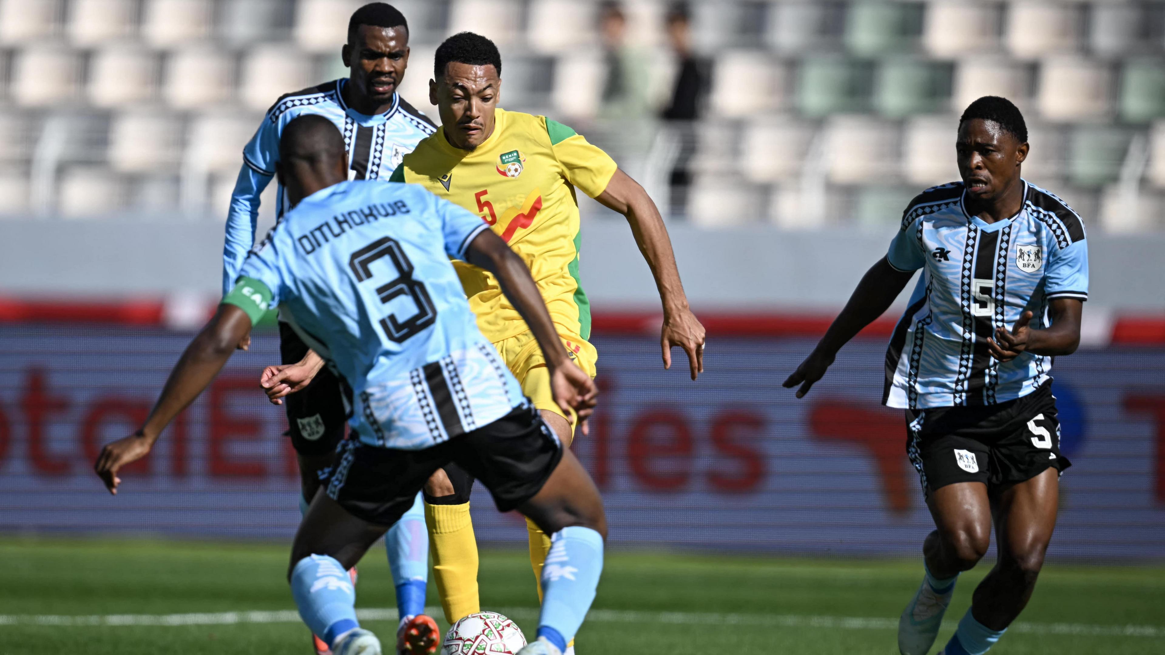 Footballer Yohan Roche takes control of the ball during the Afcon 2025 football match between Benin and Botswana, wearing a yellow, red and green kit against Botswana players in blue, black and white. Roche is seen running towards a defender with his back to the camera while two other defenders, one behind him and one to the side, attempt to close Roche down
