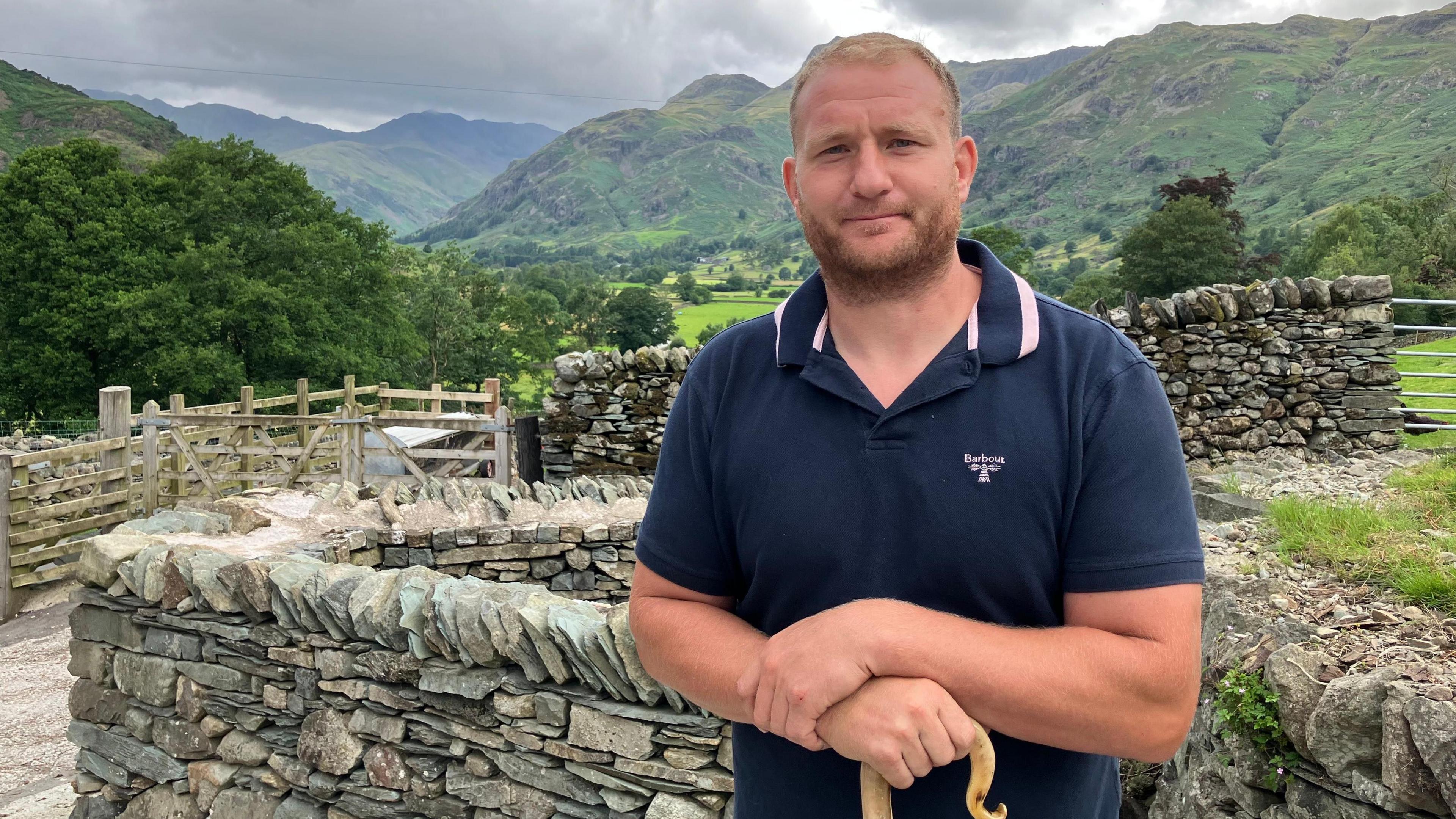 Jonathan Benson in front of a drystone wall at his farm, with the fields and fells in the background. He has short dark blond hair and a beard. He is wearing a blue polo shirt and is holding a wooden walking stick.