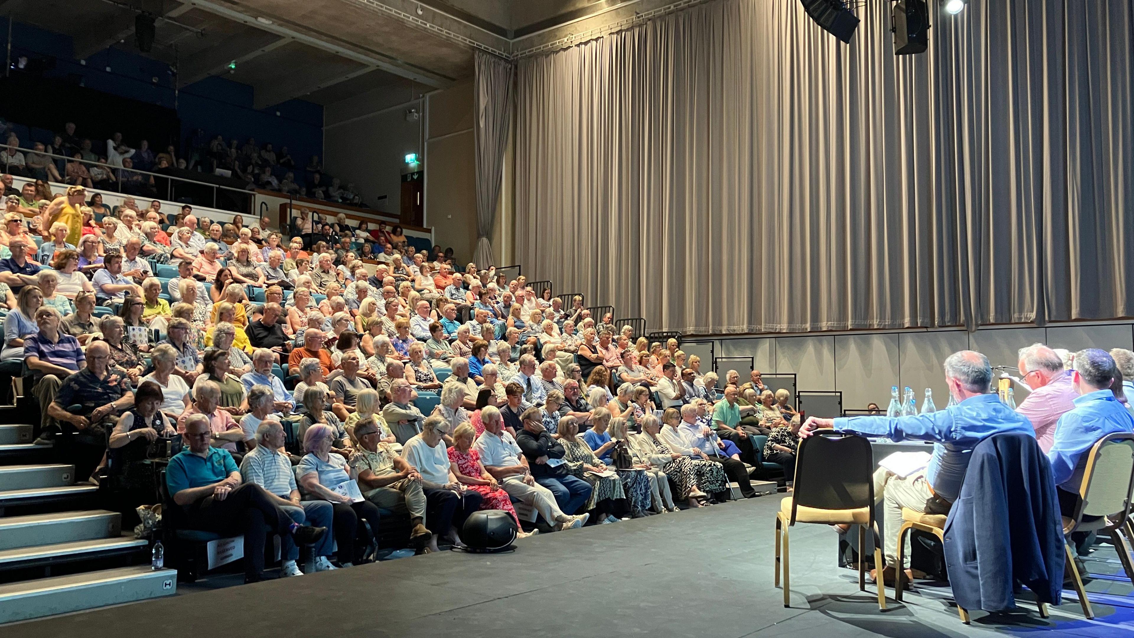 Auditorium style room with hundreds of people sat in a tiered seating area looking at panel of men in shirts.