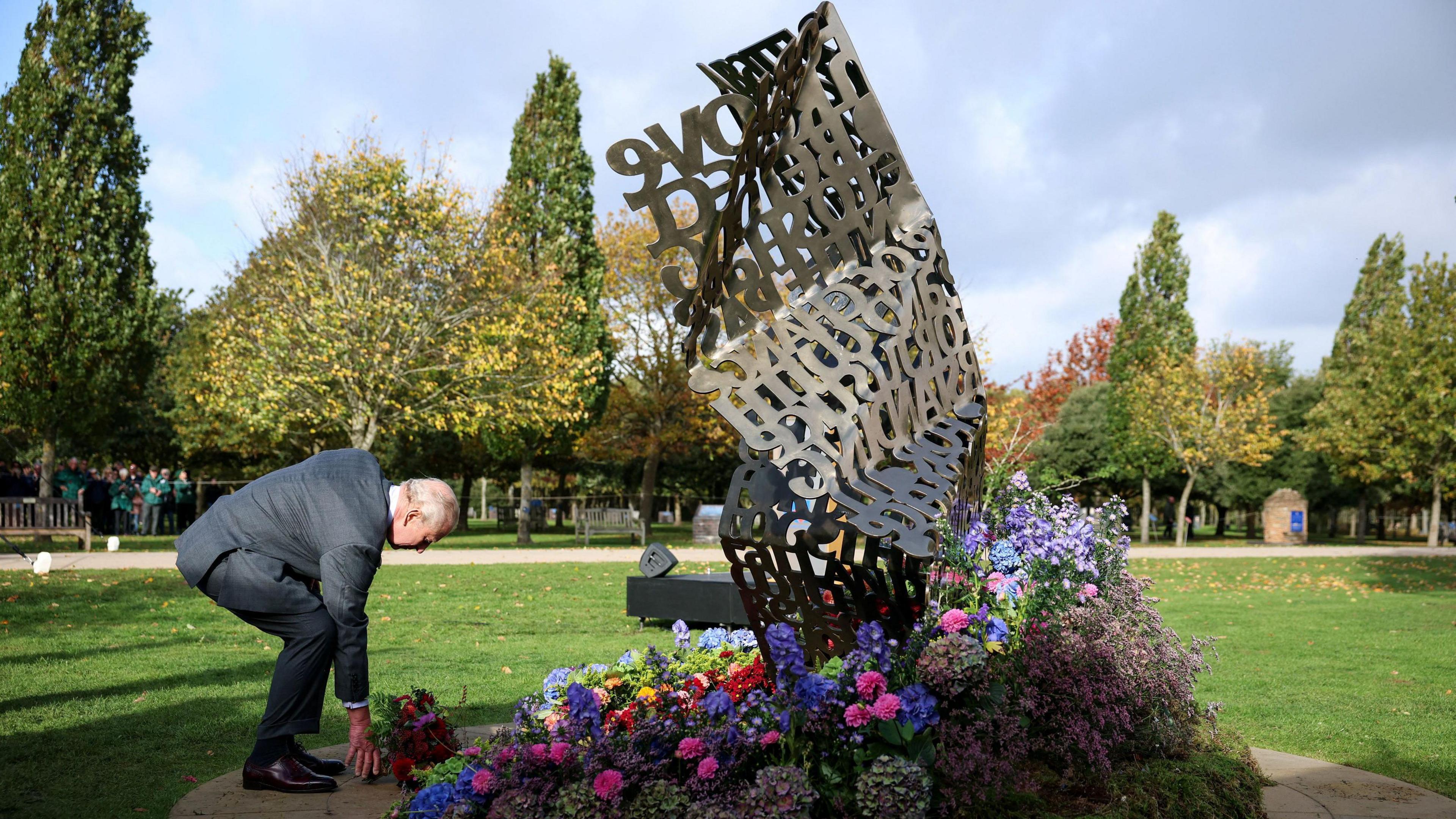 King Charles, wearing a grey suit, places flowers down by a large grey sculpture that resembles a crumpled piece of paper made up of words. There are grass and trees in the background.
