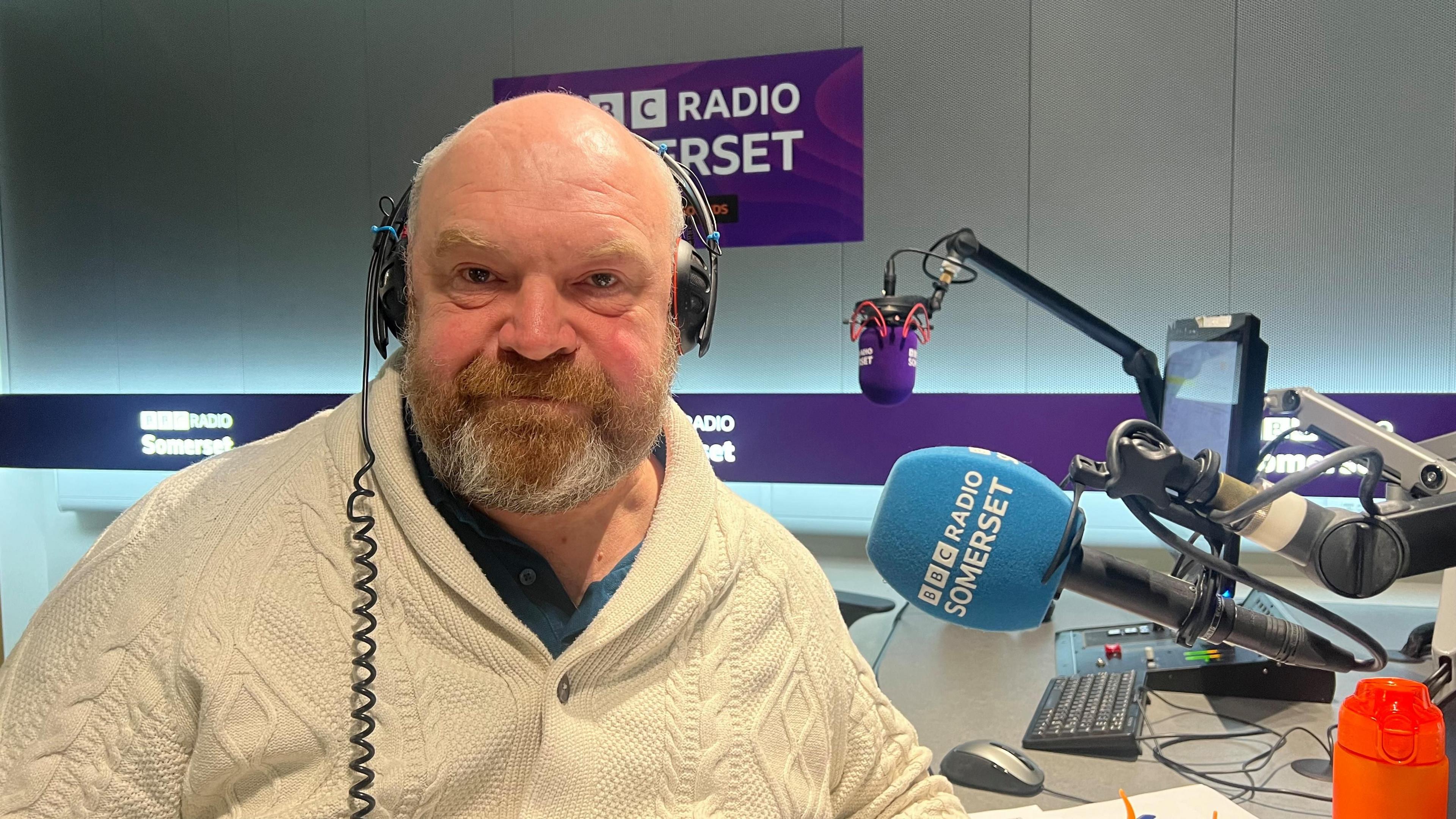 A man in a cream cardigan sitting in front of a microphone in a radio studio