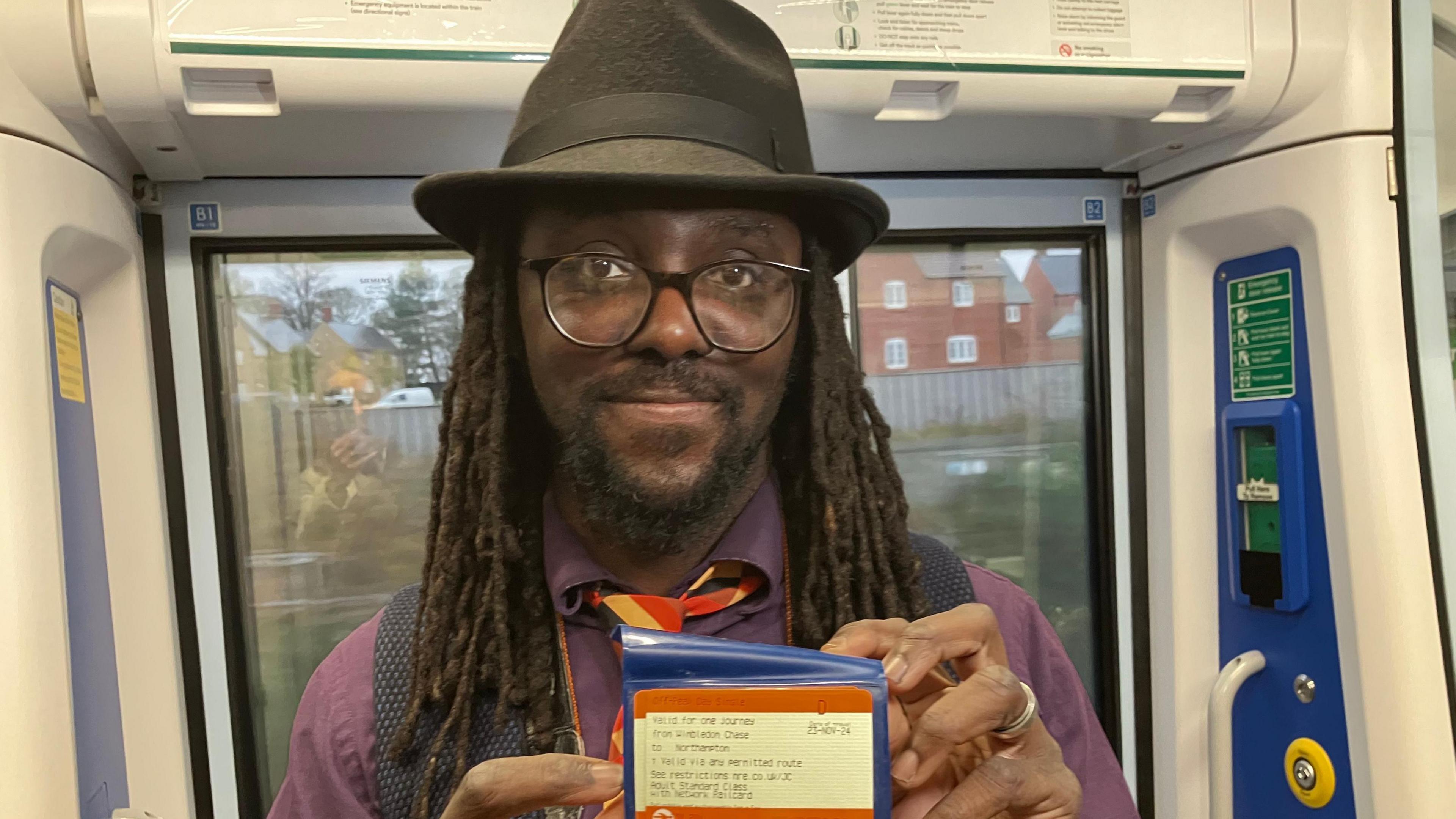 Luke Agbaimoni holding up a train ticket. He is wearing glasses, a trilby hat and a purple shirt under a black waistcoat and yellow and orange tie. He has dark dreadlocks falling over his shoulders 