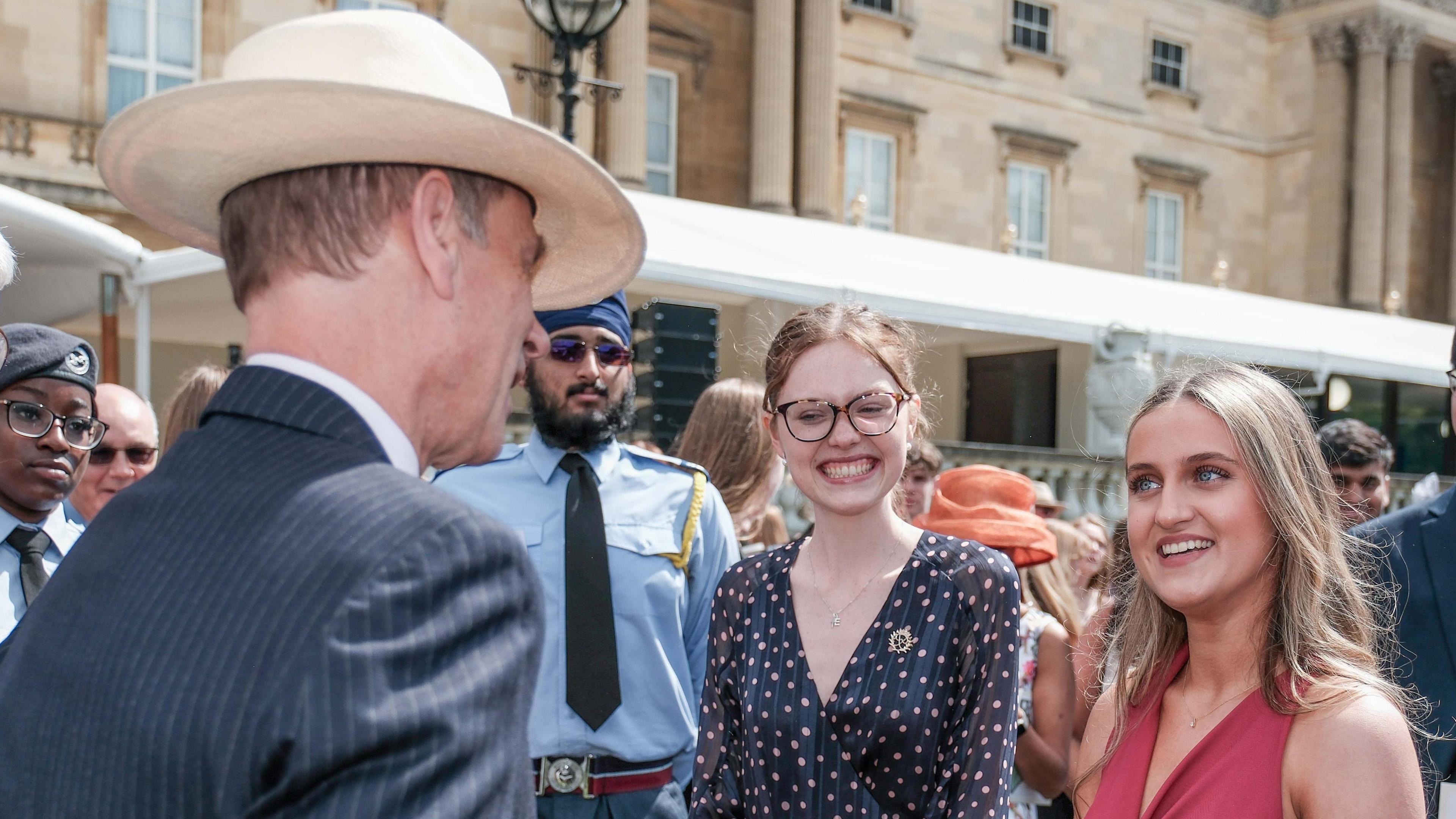 The Duke of Edinburgh, talking to Lucy, at a special garden party, with several people around her. The Duke is wearing a suit pinstriped suit, a hat and has his back to the camera. Zoe is smiling and looking at him.