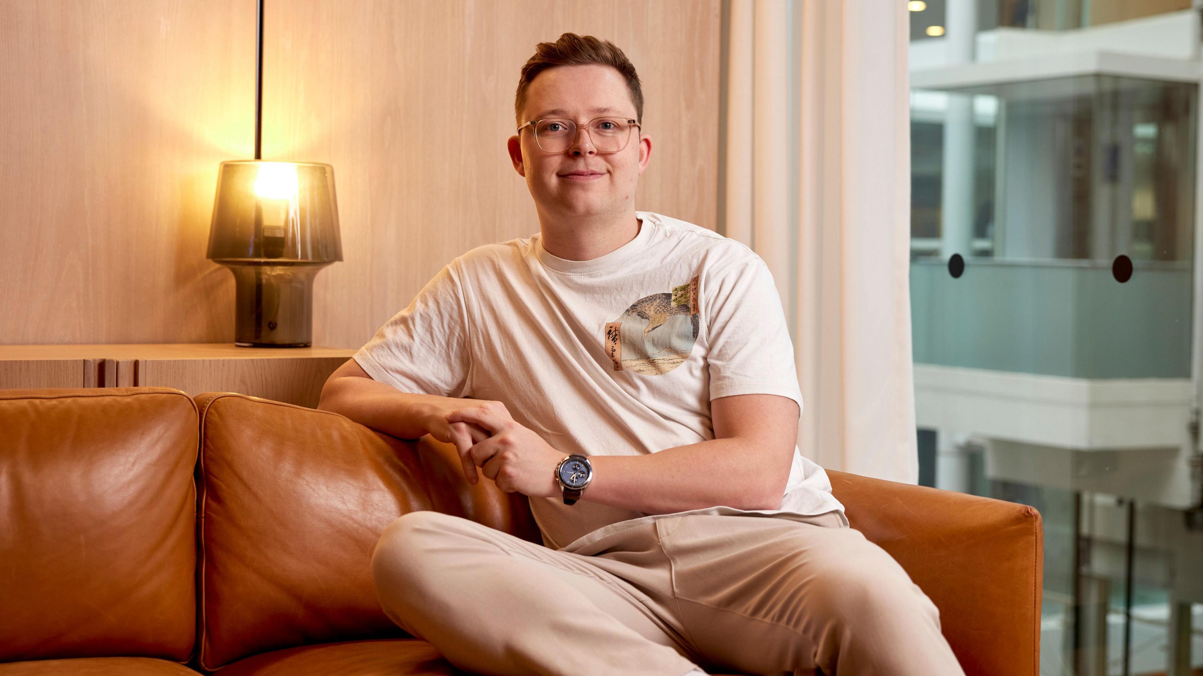 James Courtney sits on a light brown leather sofa and smiles at the camera - he wears a white t shirt and beige trousers with glasses and a watch