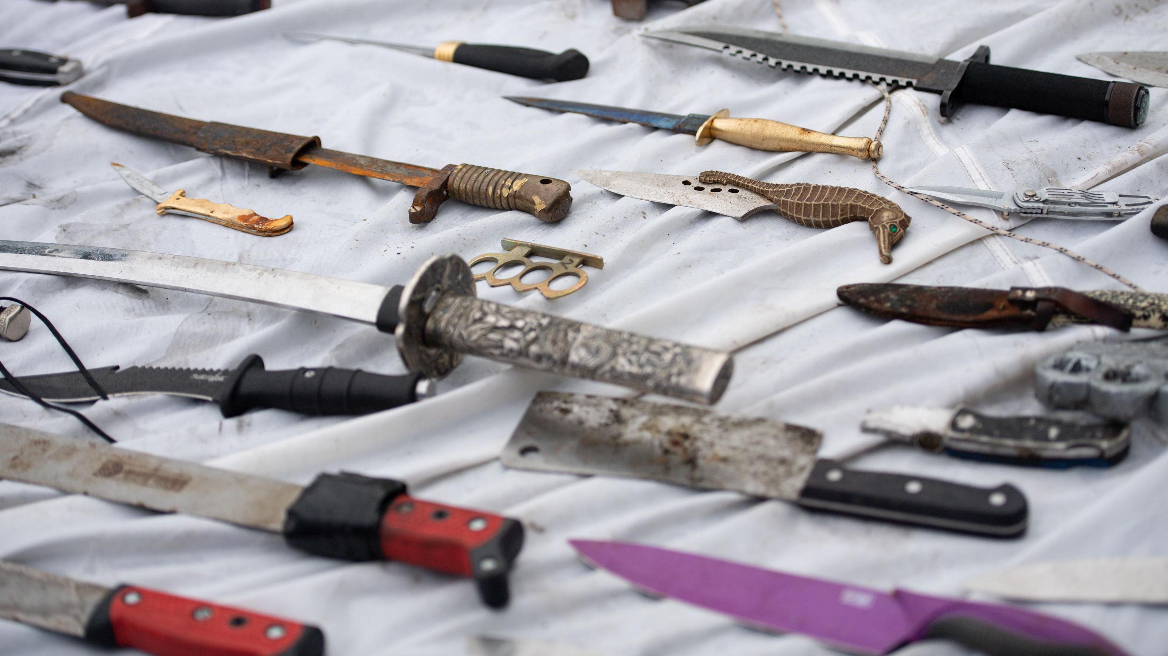 Knives and other weapons laid on a white sheet that have been deposited into weapon surrender bins.