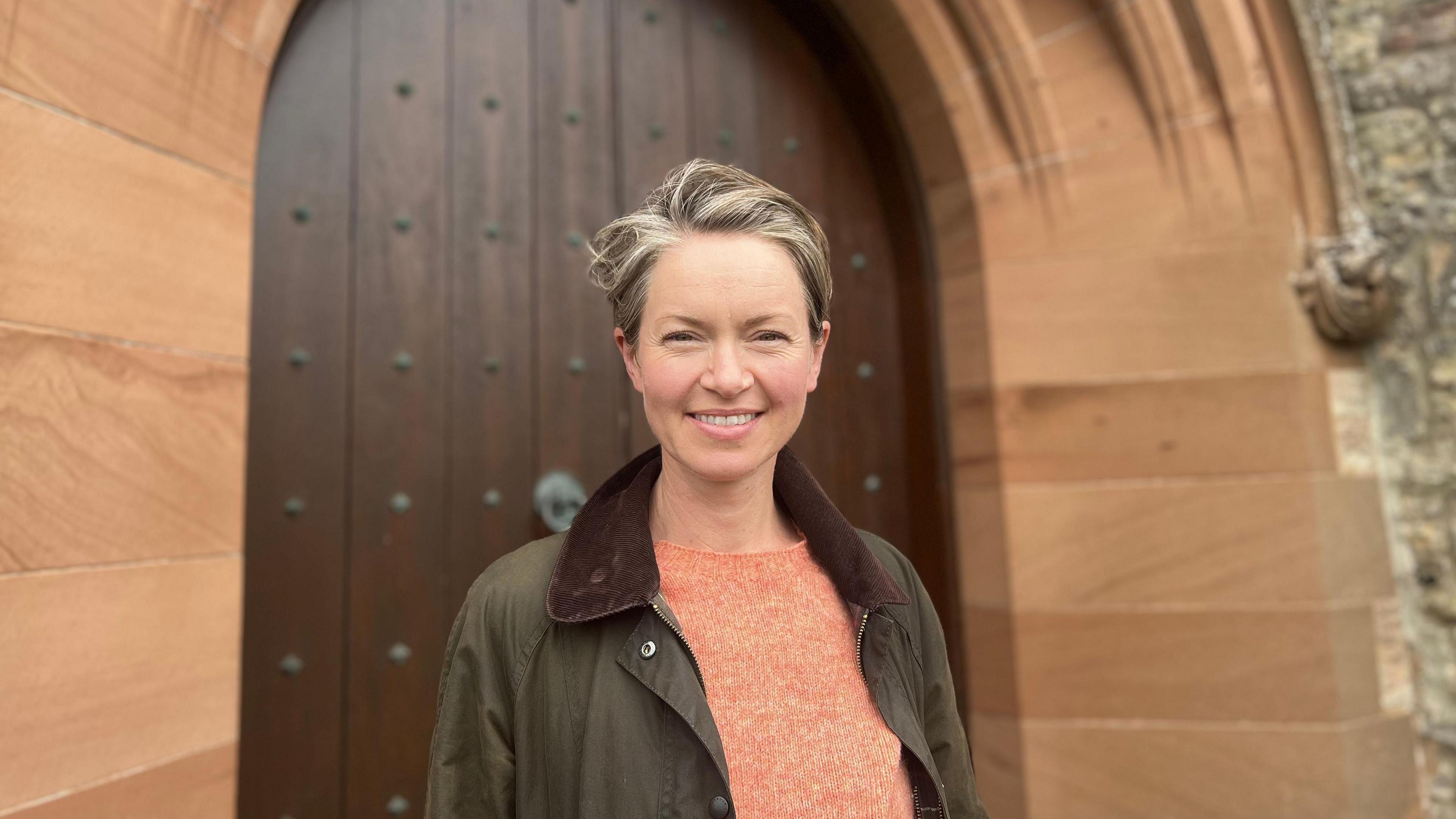 A woman with short brown hair, wearing an orange top and brown jacket, stands in front of a church door