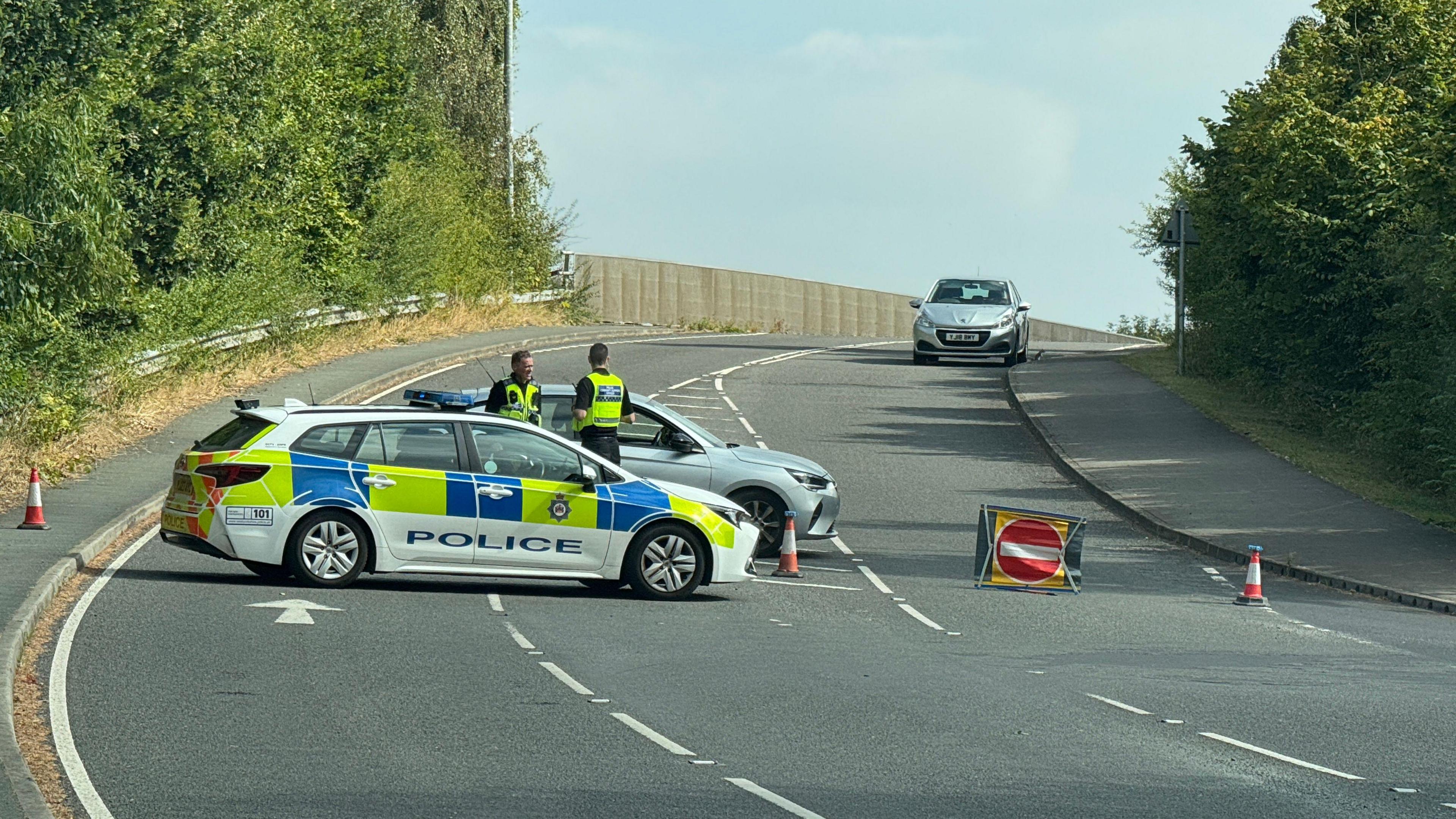 A police car is parked across two lanes on York Road. Two police officers are stood behind the vehicle talking and there is a no entry sign and traffic cones blocking off the road.