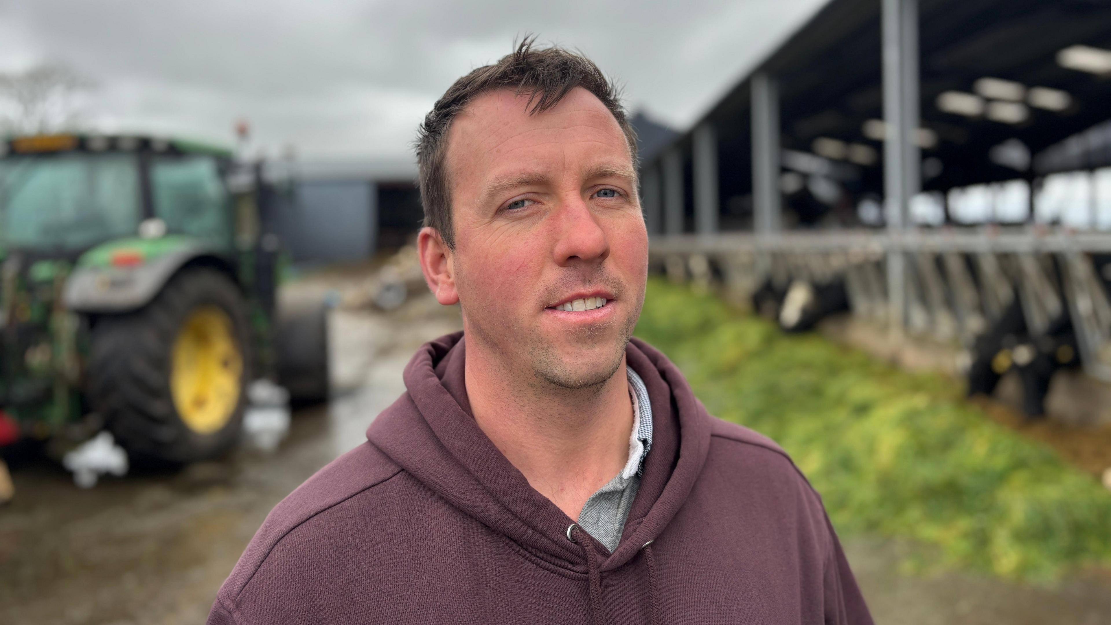 A young farmer stands in his farmyard. He's dressed warmly in a plum colored hoody he has short hair and a neutral expression. behind him. is agreen tractor and a shed of cows. The cows are assembled along the edge of the shed to eat the bright green grass lying outside.