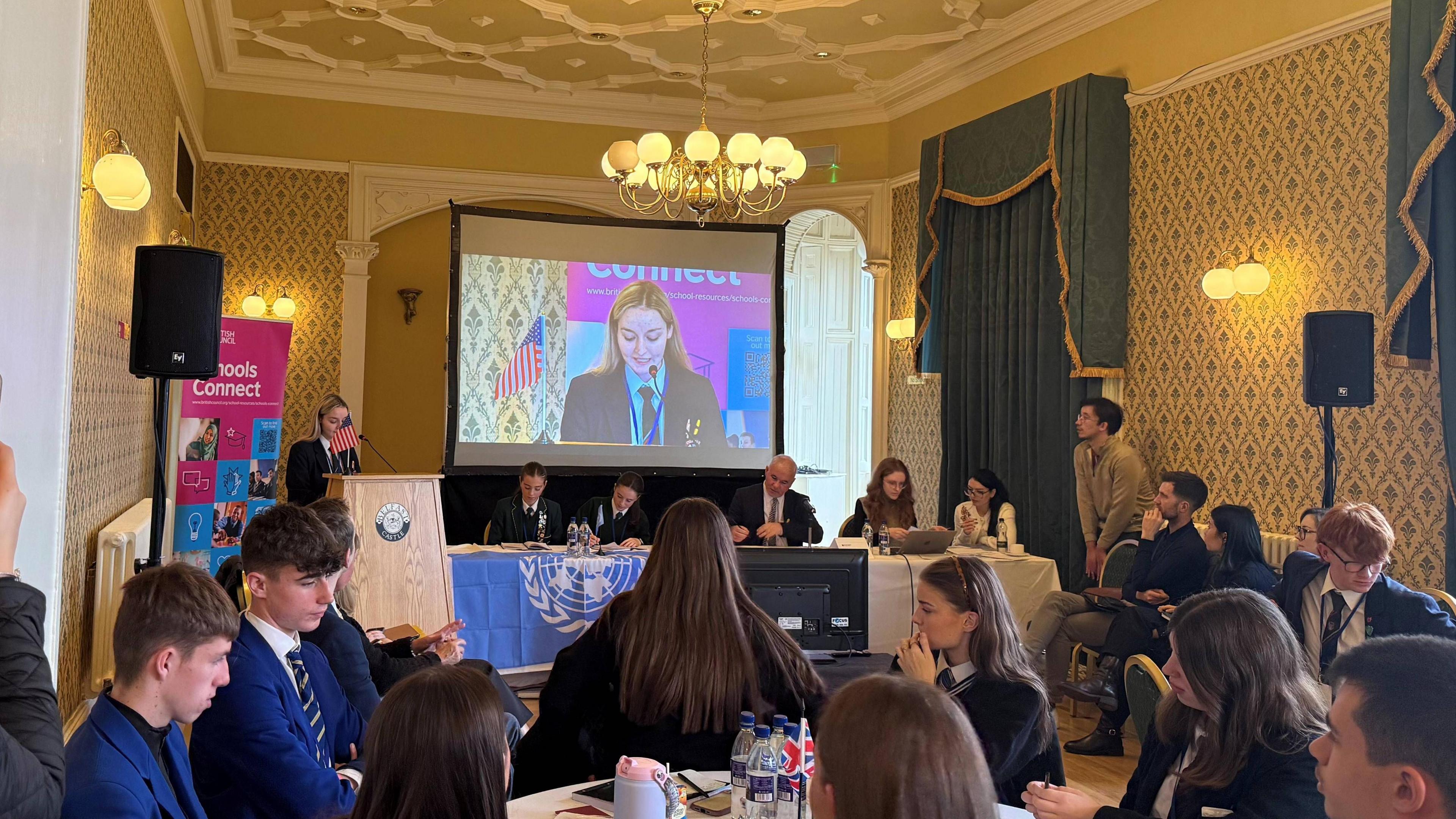 Around 20 people are sitting at tables in a conference room. One girl is standing at a podium. She is being shown on a projector screen in the middle of the room.