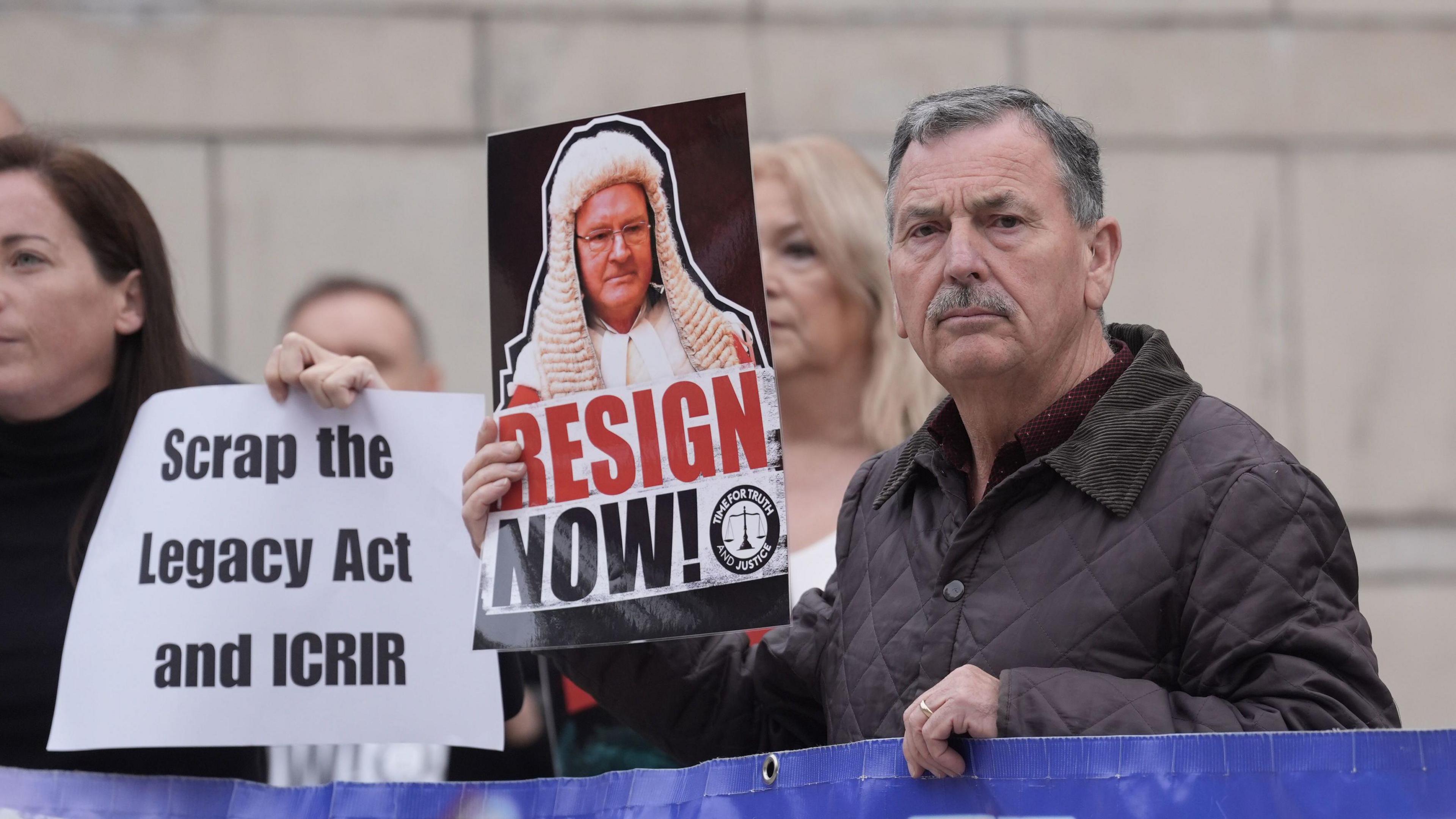 John Teggart, the son of Daniel Teggart who was killed in the Ballymurphy massacre in 1971, outside the Court of Appeal at the Royal Courts of Justice in Belfast. He is holding a picture of Sir Declan Morgan saying 'Resign Now'. Another sign is held in the background saying: 'Scrap the Legacy Act and ICRIR'