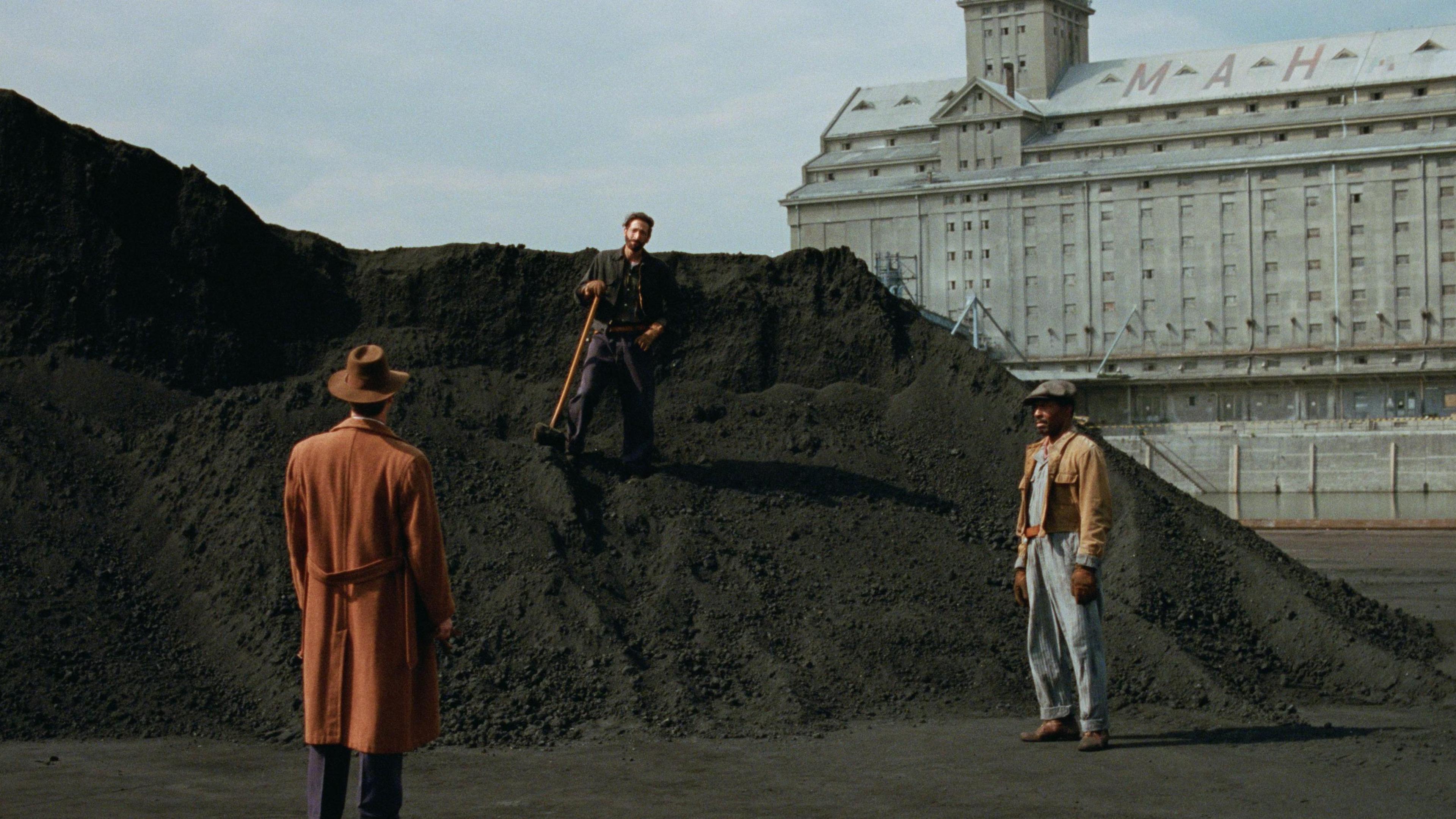 A scene from The Brutalist featuring three men. One man, who has dark hair and clothes, is stood on a large pile of rubble. In front of him are two other men, one in a brown hat and coat with his back to the camera, and the other in a casual brown jacket and blue jeans and flat cap. Behind them is a grand grey building with the letters MAH on its roof.