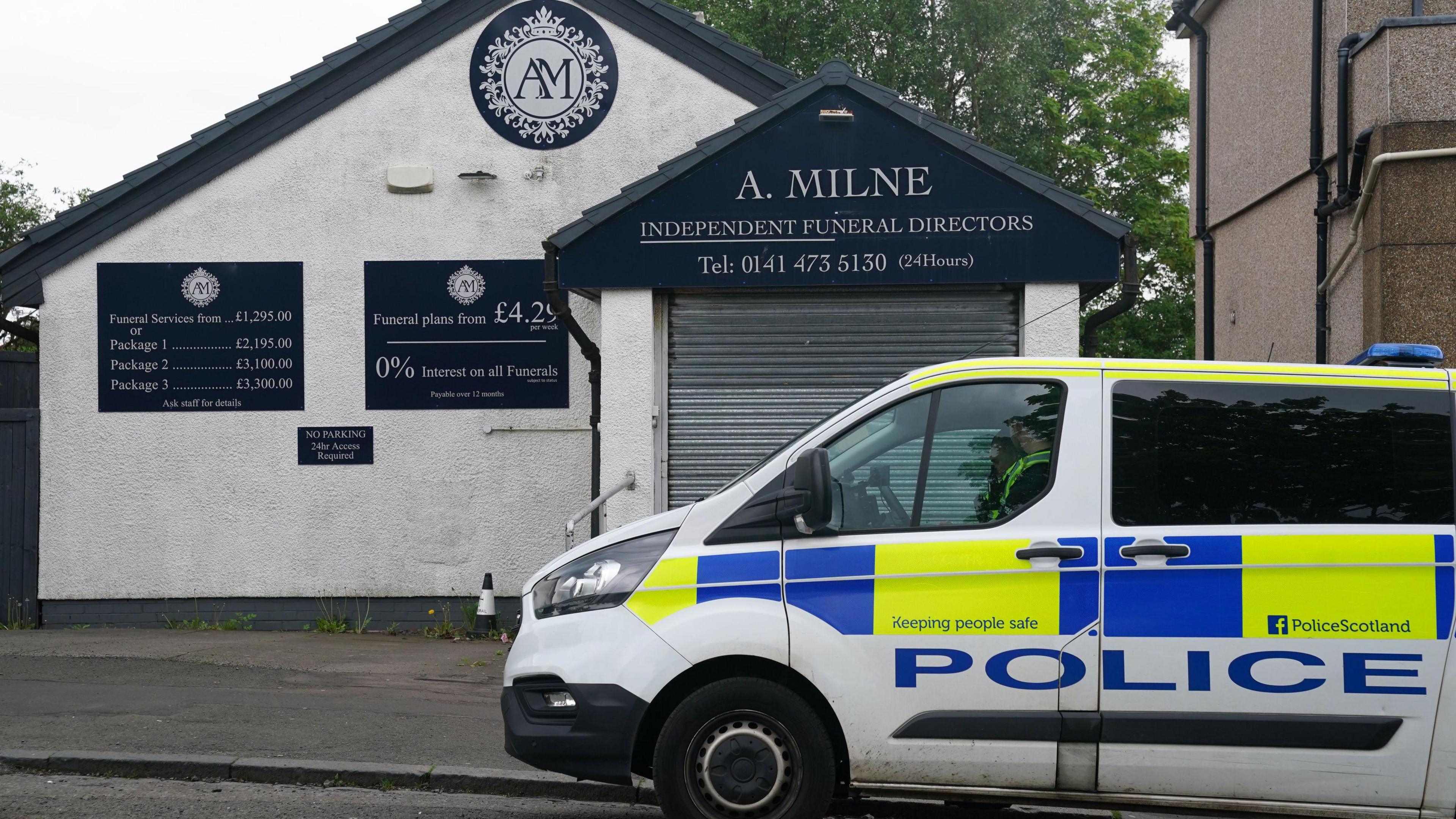 A police van parked outside a white building which has its front shutter down. There are signs on the building saying A Milne Funeral directors