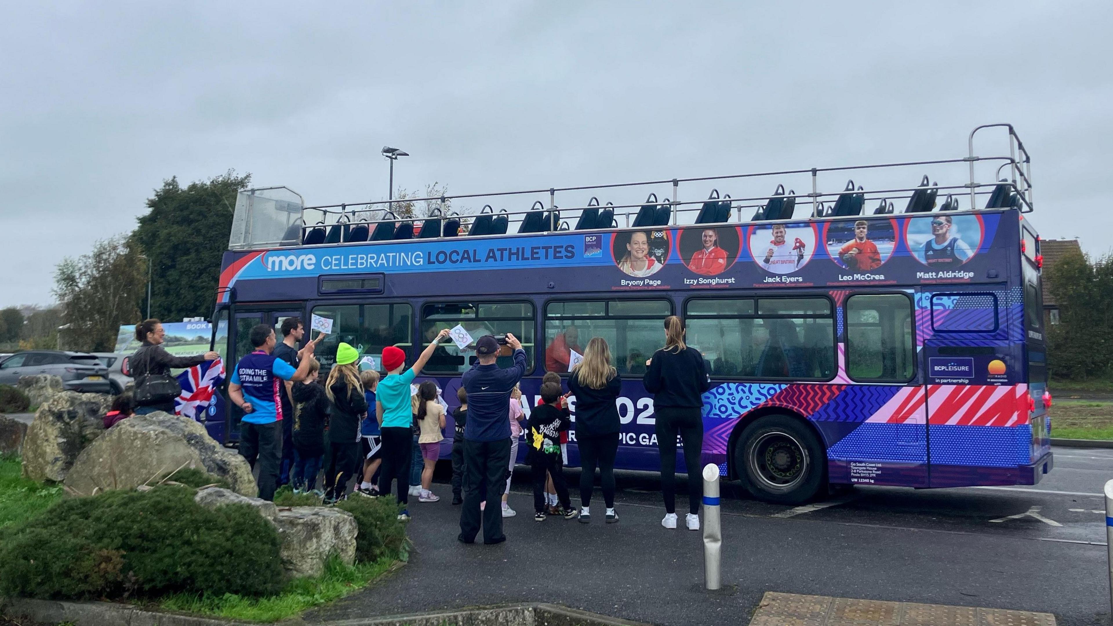 A purple double decker bus with the athletes' faces on, with a small crowd of people standing next to it, some waving flags.