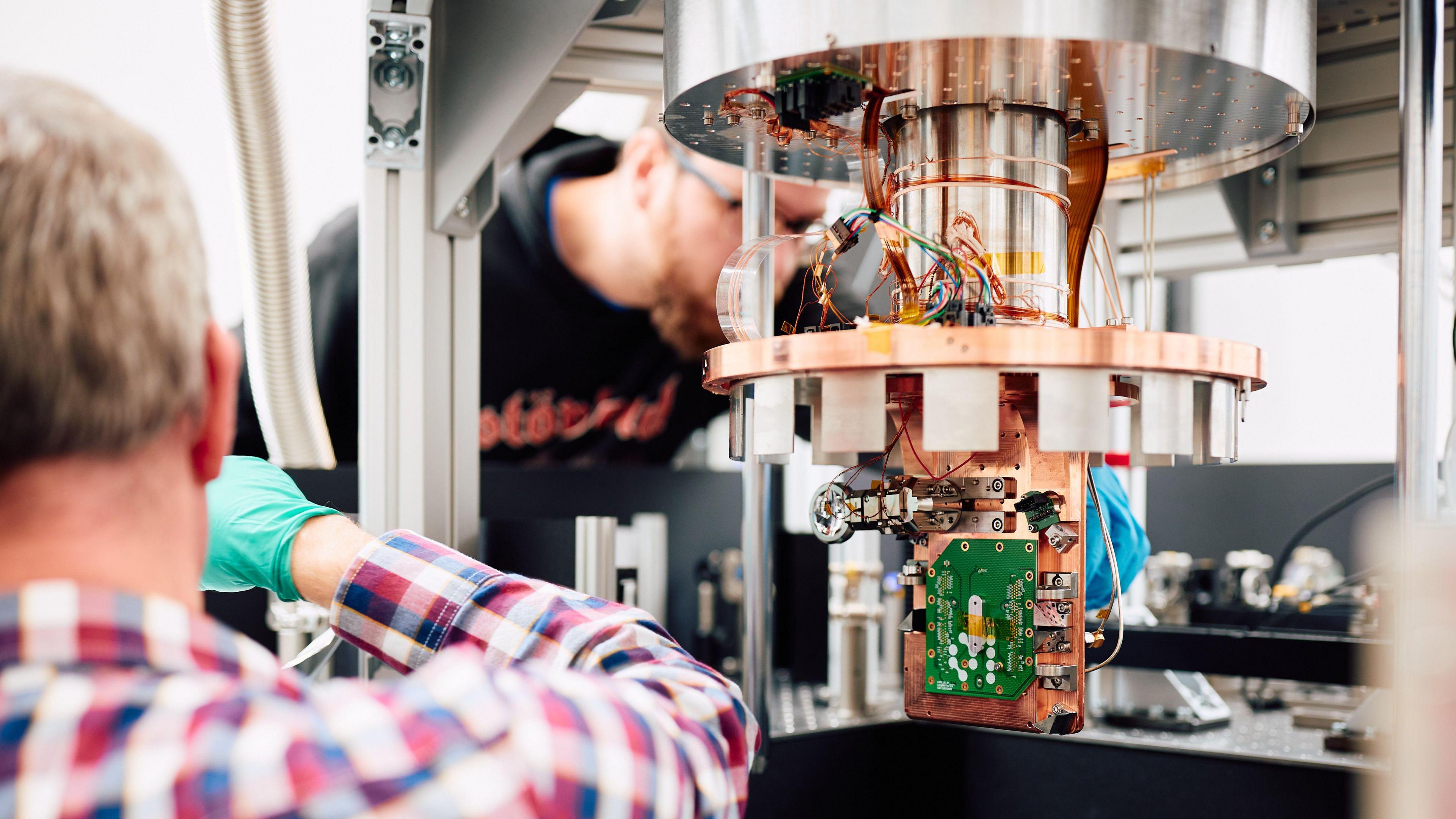 A green computer chip attached to a large copper coloured fixture with two men, both wearing gloves, working on it