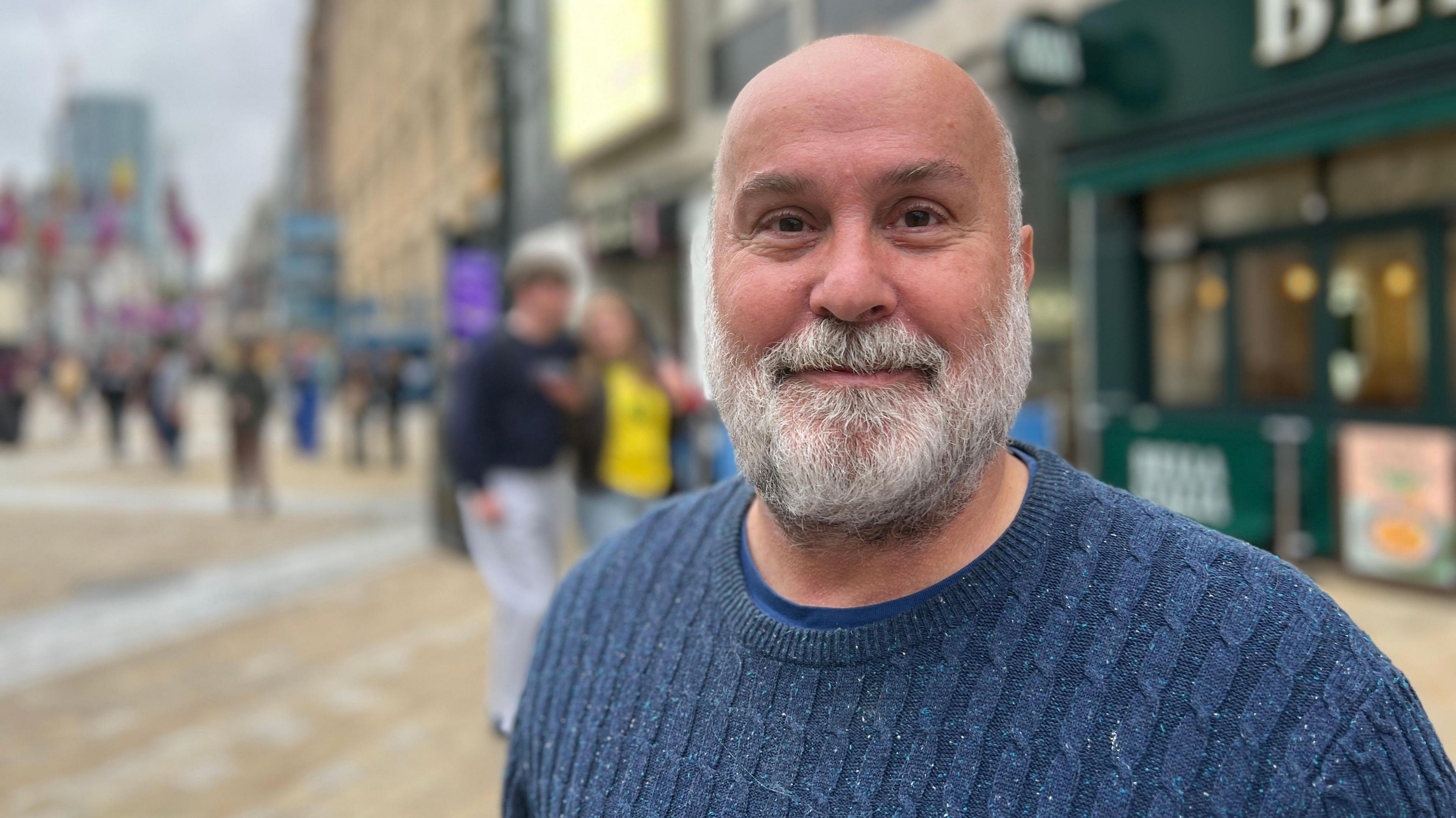 A man with a grey beard is in centre frame. He wears a blue jumper. Behind him, Briggate is blurred.